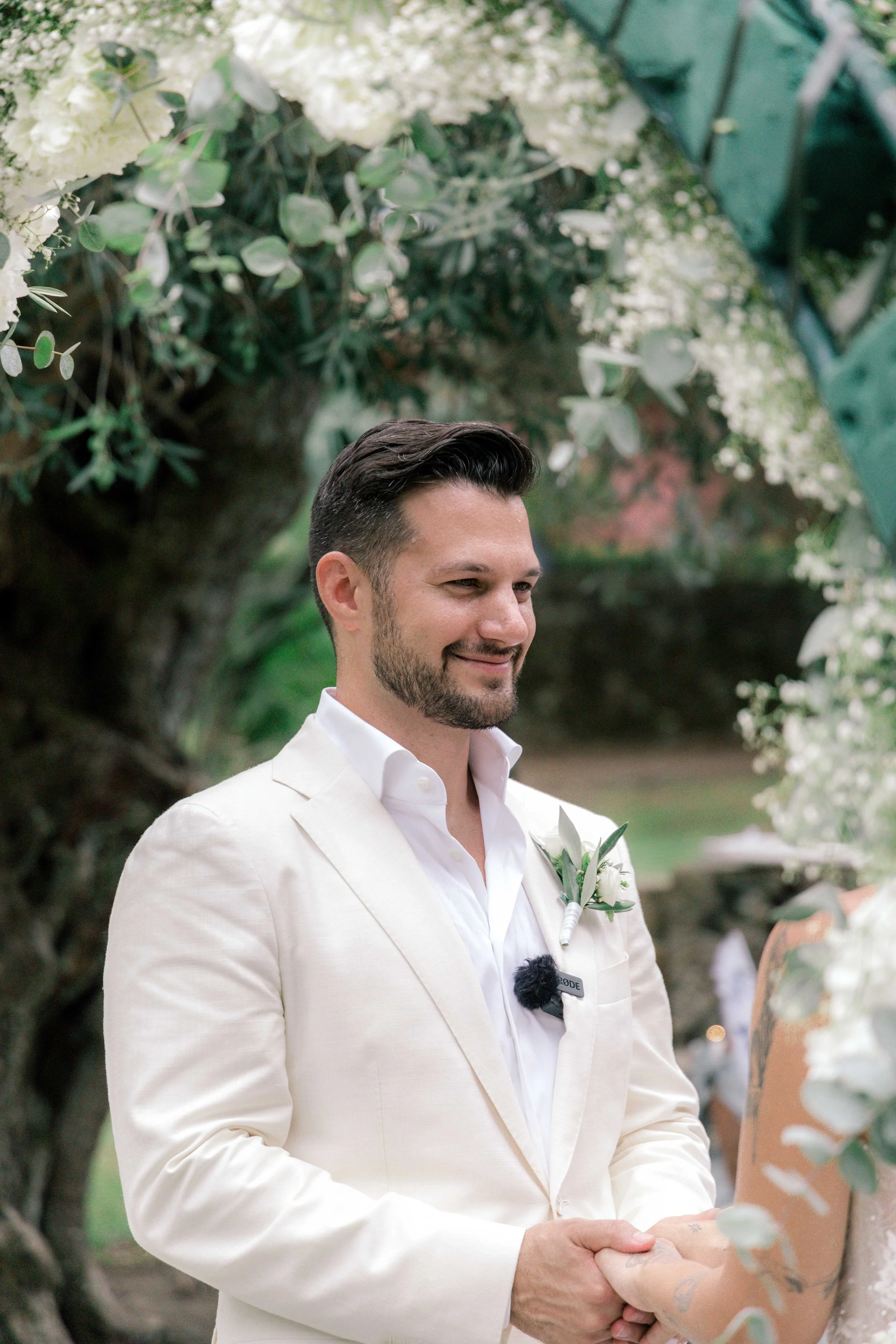 A groom in a light-colored blazer with a boutonniere, smiling during his wedding ceremony outdoors under a floral arch.