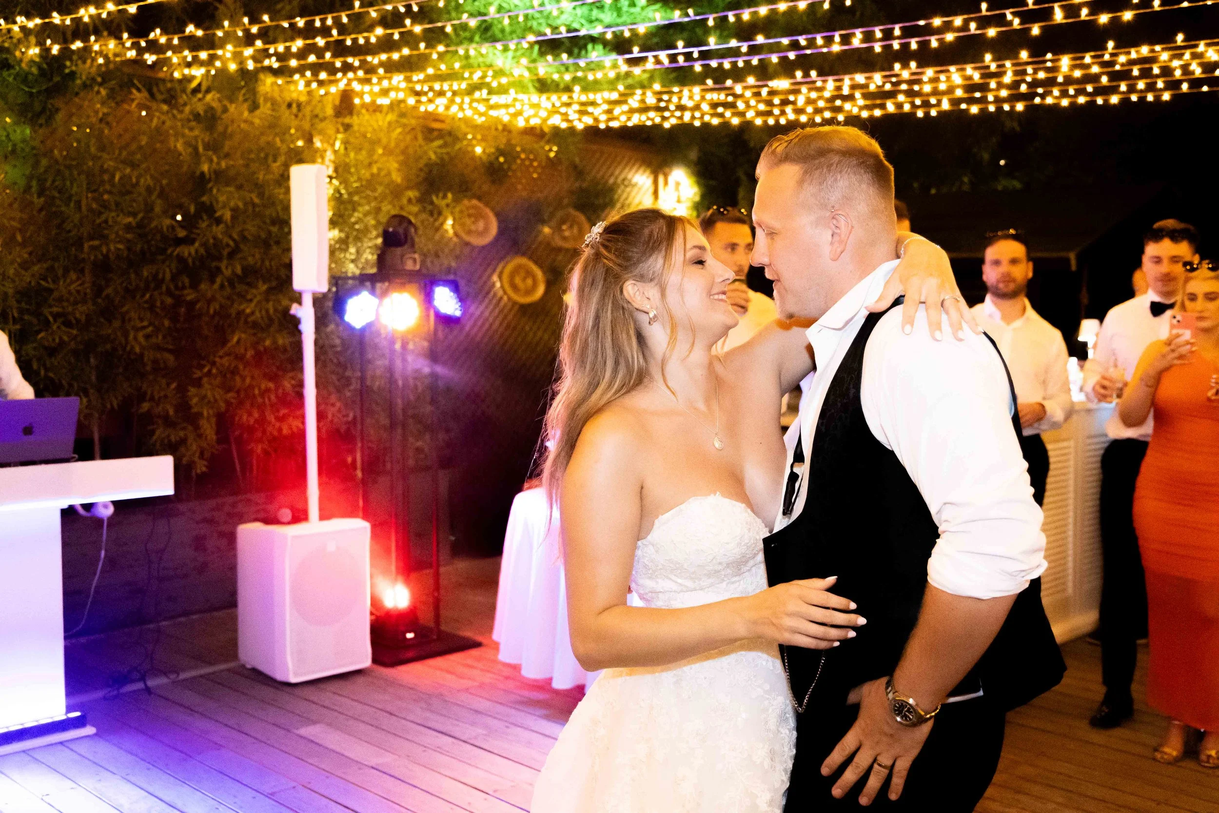 A bride and groom dance closely at their wedding reception with guests watching in the background. String lights hang overhead, creating a festive atmosphere.