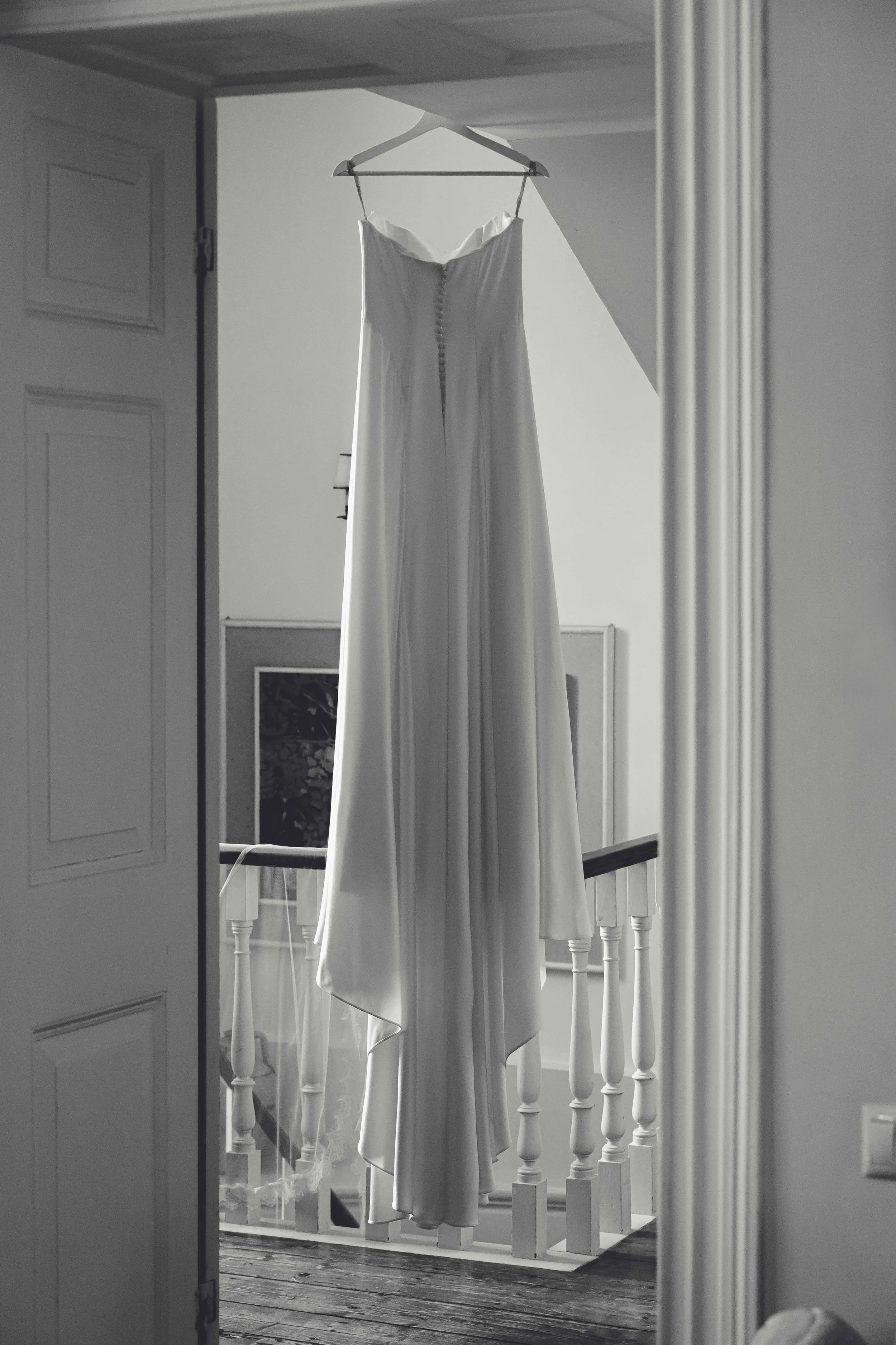 A white wedding dress hanging from a hanger on a wooden beam inside a house, with a staircase and a white railing in the background.