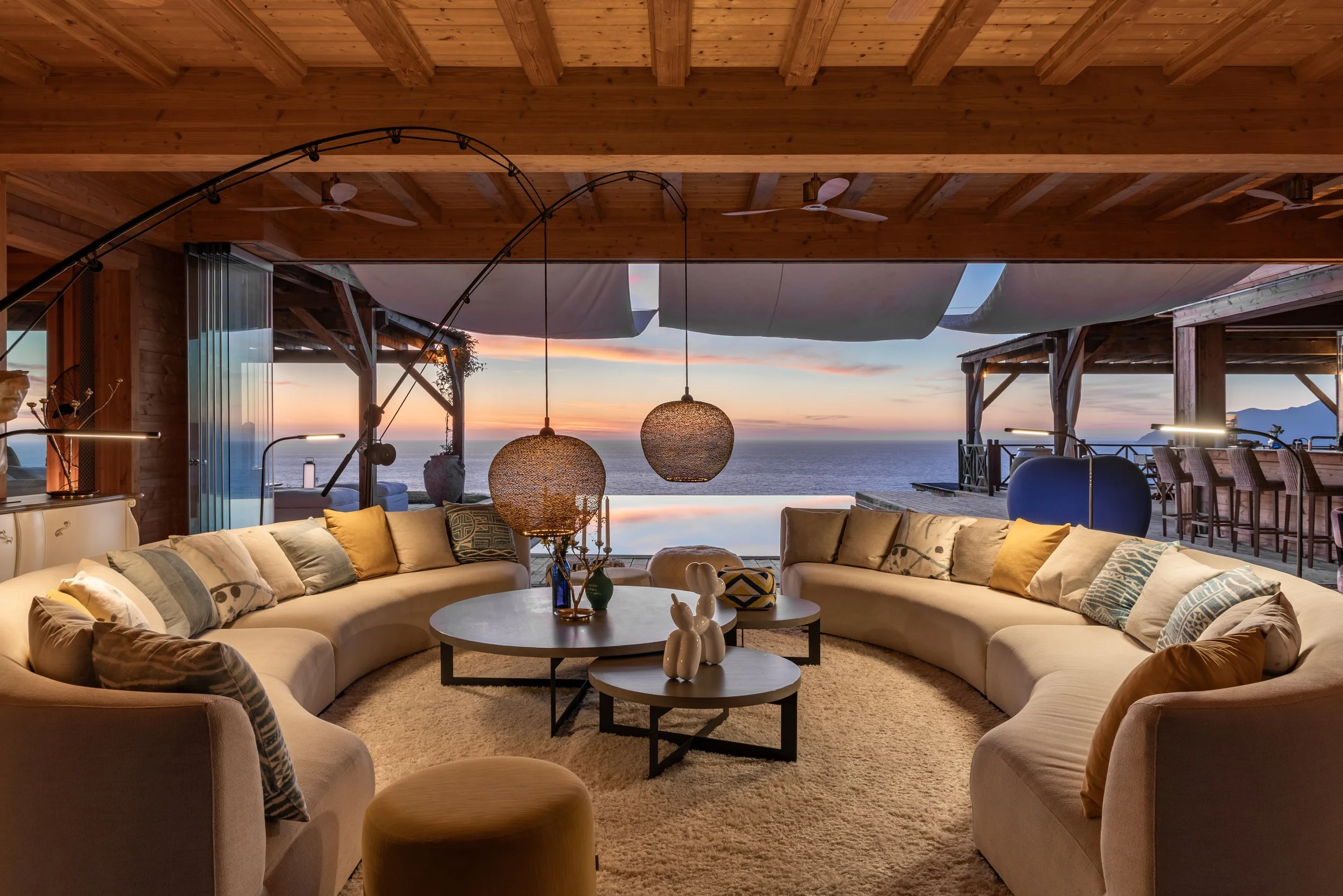 Living room with large curved sofa, coffee tables, pendant lights, and ocean view at sunset