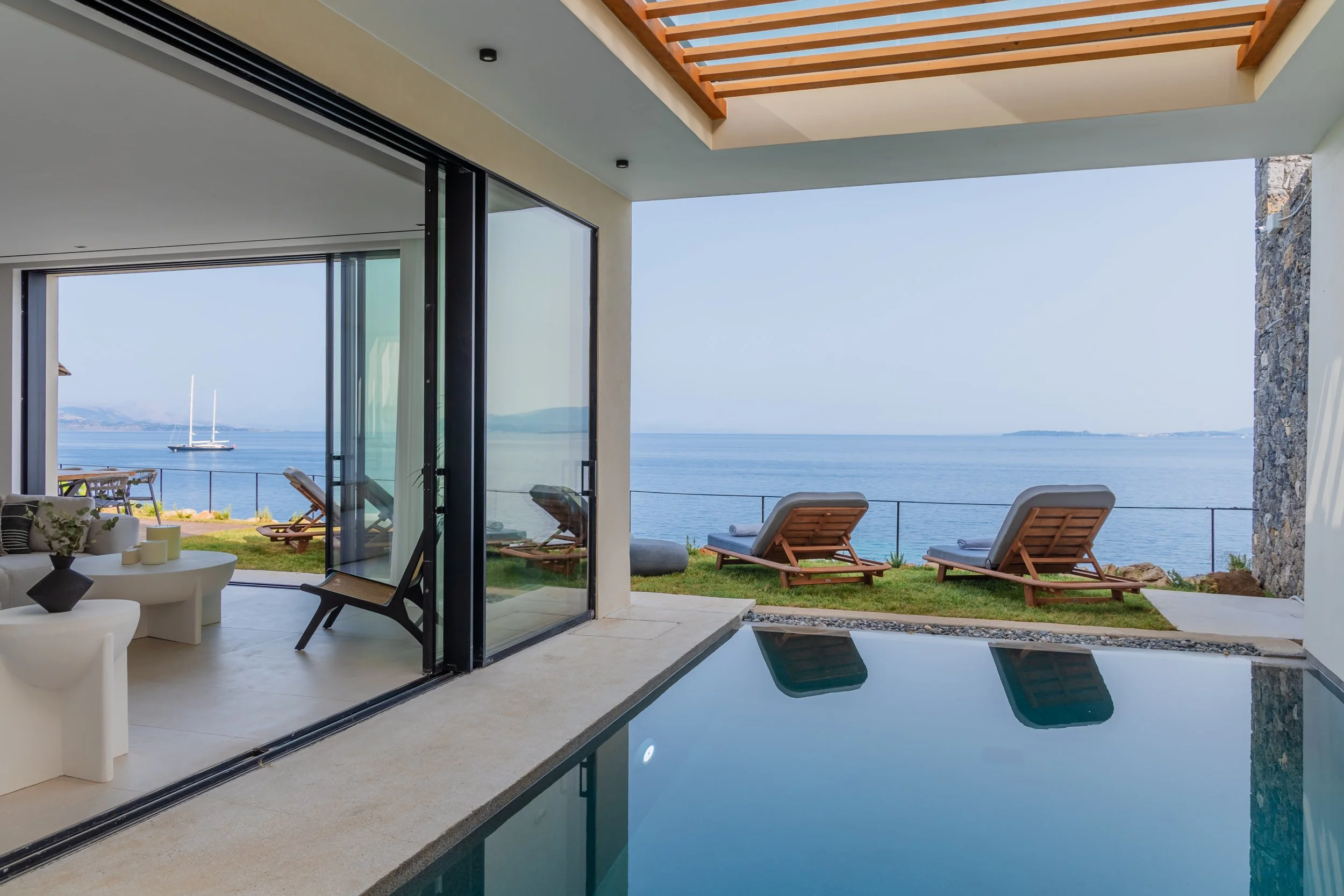 View of a modern house with large glass doors opening to a patio with lounge chairs and a view of the ocean and distant islands.