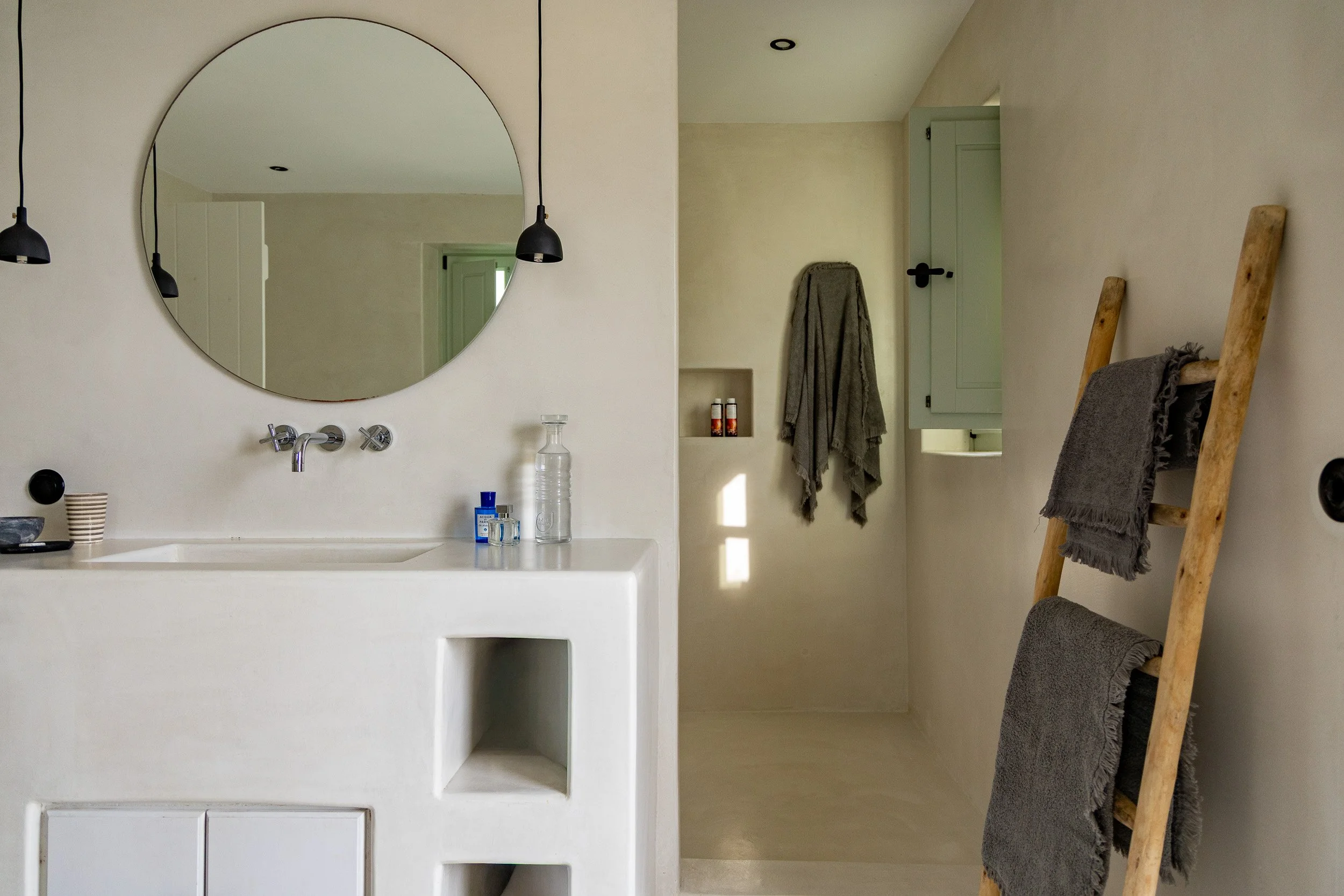 Minimalist bathroom with a white vanity, large round mirror, black pendant lights, and rustic ladder with gray towels.