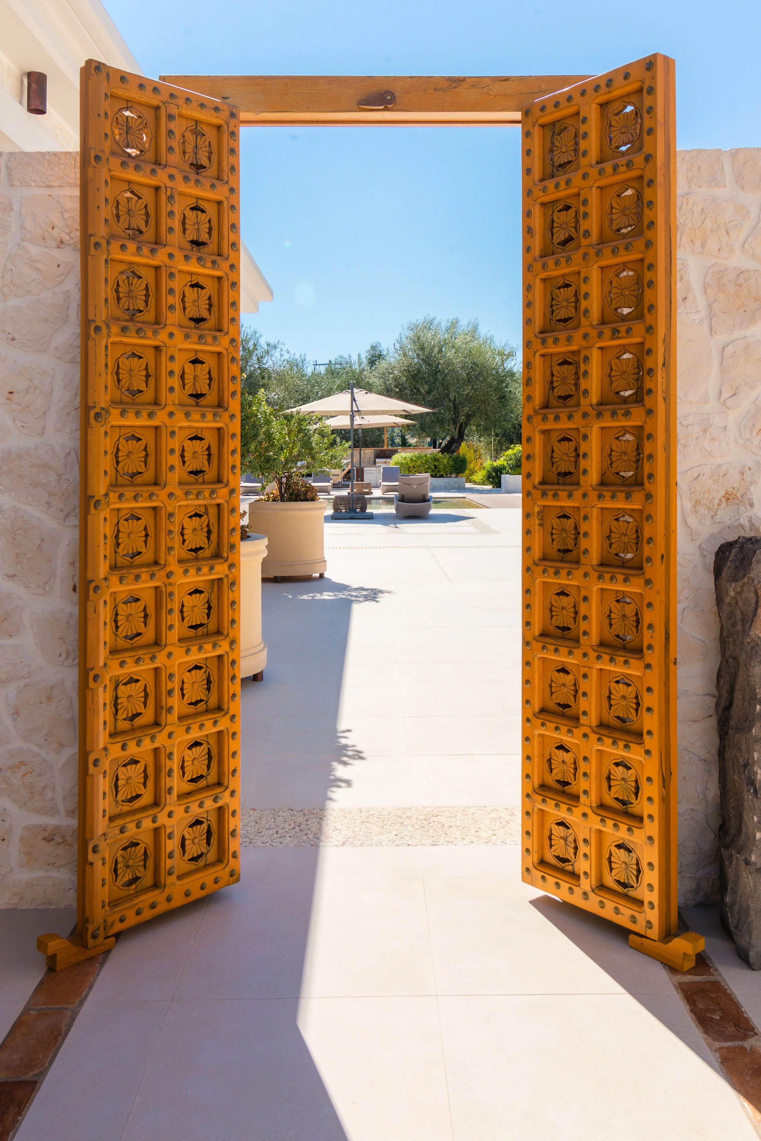 Open wooden doors revealing a patio with outdoor seating, umbrellas, and trees under a clear blue sky.