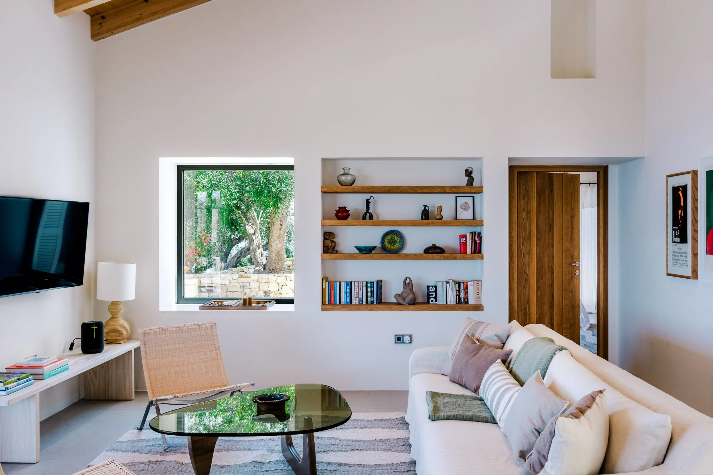 Living room with white walls, a large window showing a tree outside, a flat-screen TV on the left, a small side table with a lamp, and a beige sofa with multiple pillows. There is a wooden door on the right and built-in wooden shelves with decorative