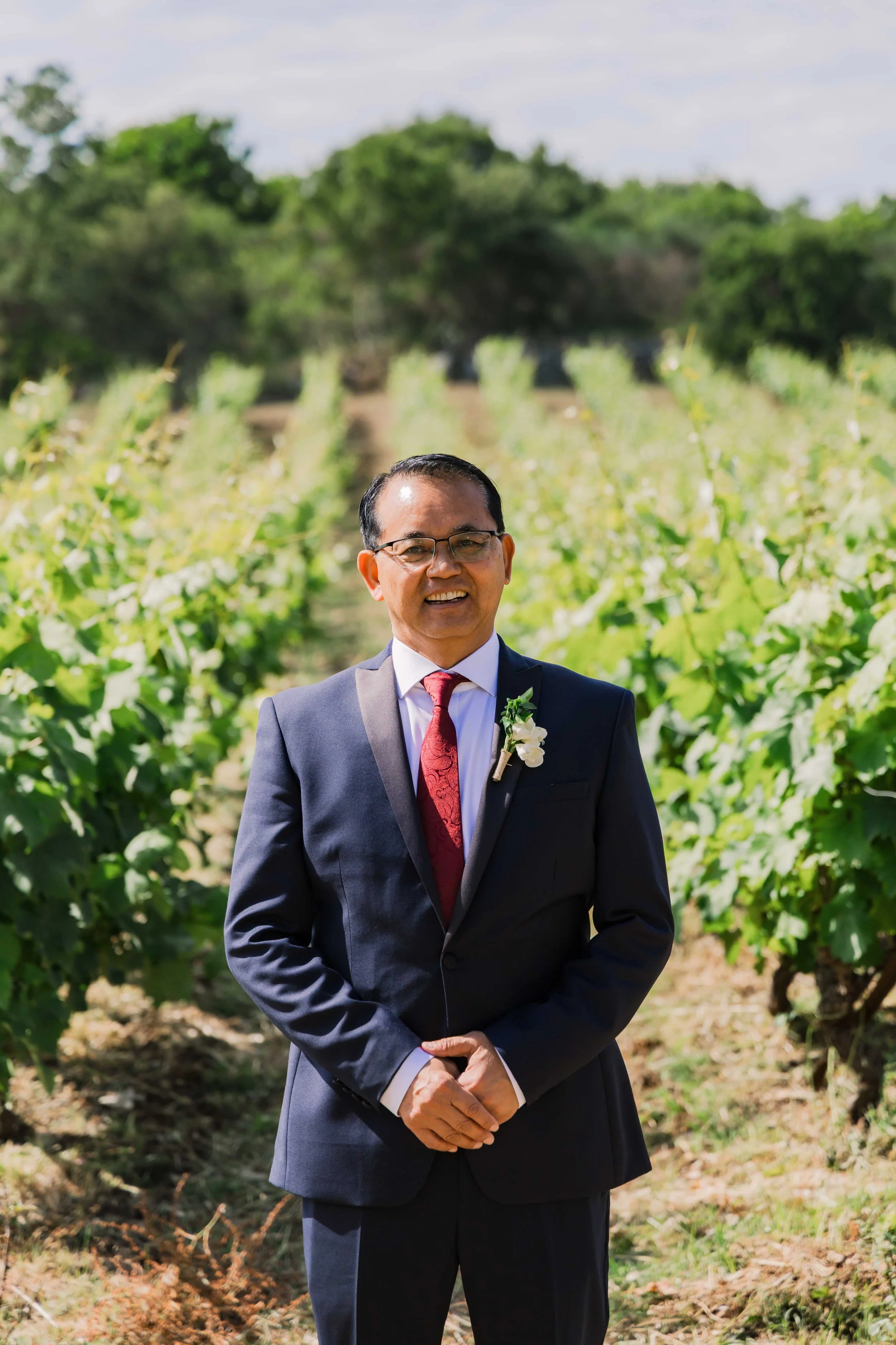 A man dressed in a suit and red tie stands in a vineyard, smiling.