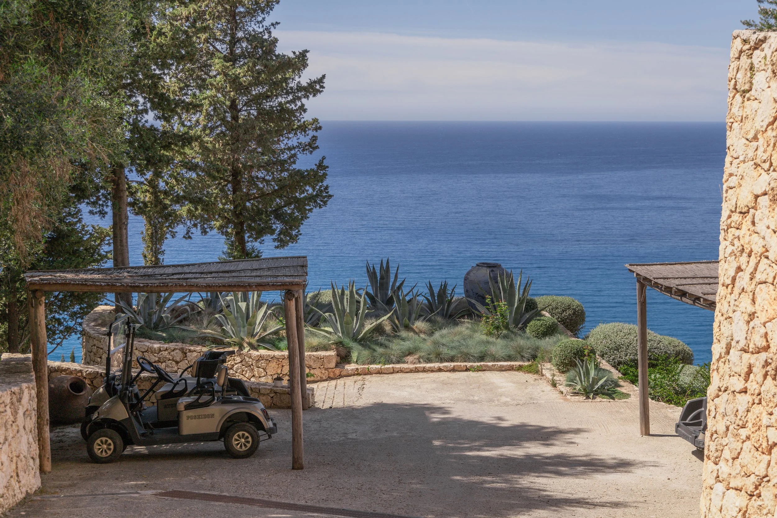 Golf carts parked under wooden shelters on a patio, overlooking a garden with succulents and the ocean beyond.