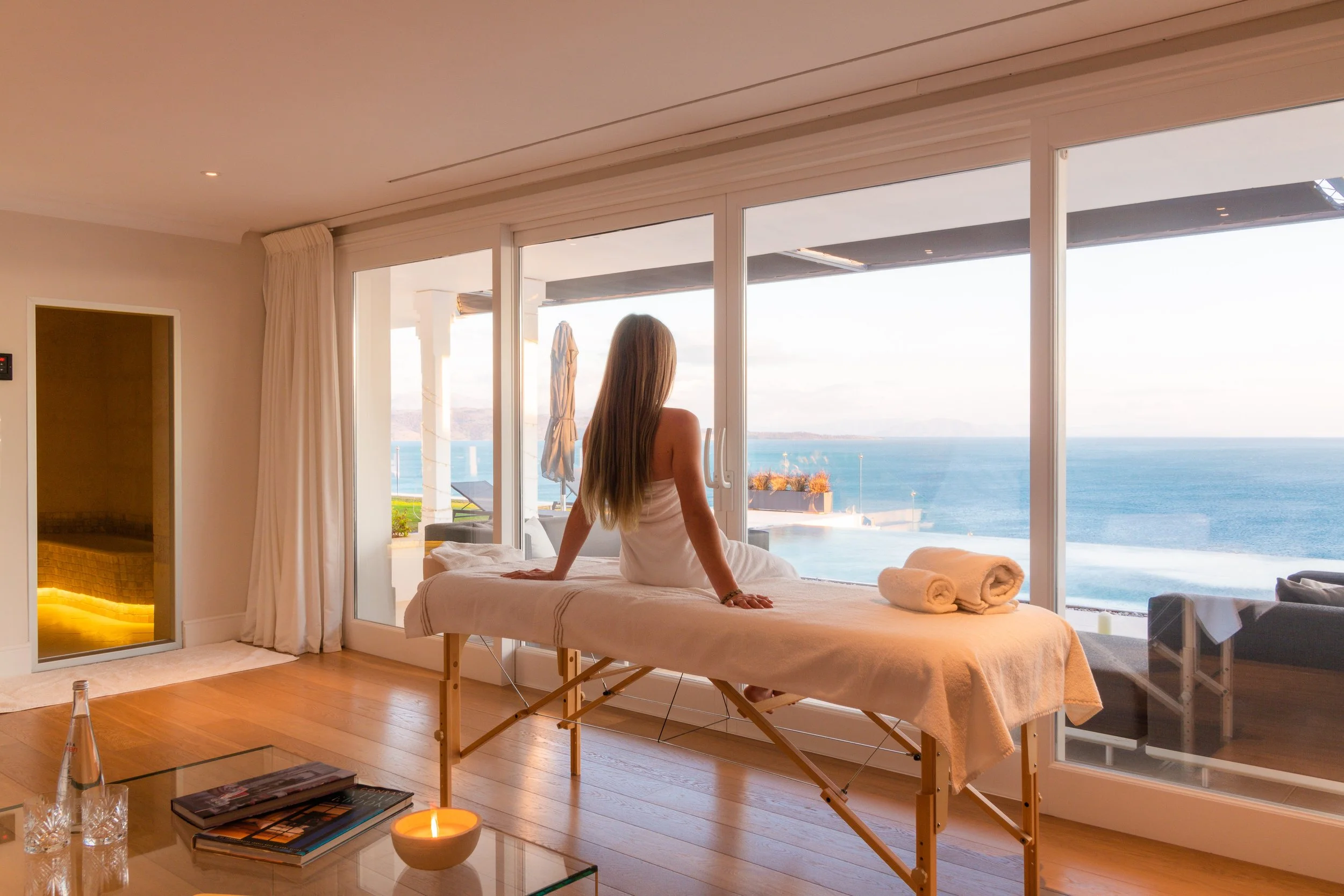 Woman sitting on massage table overlooking ocean view through large glass doors