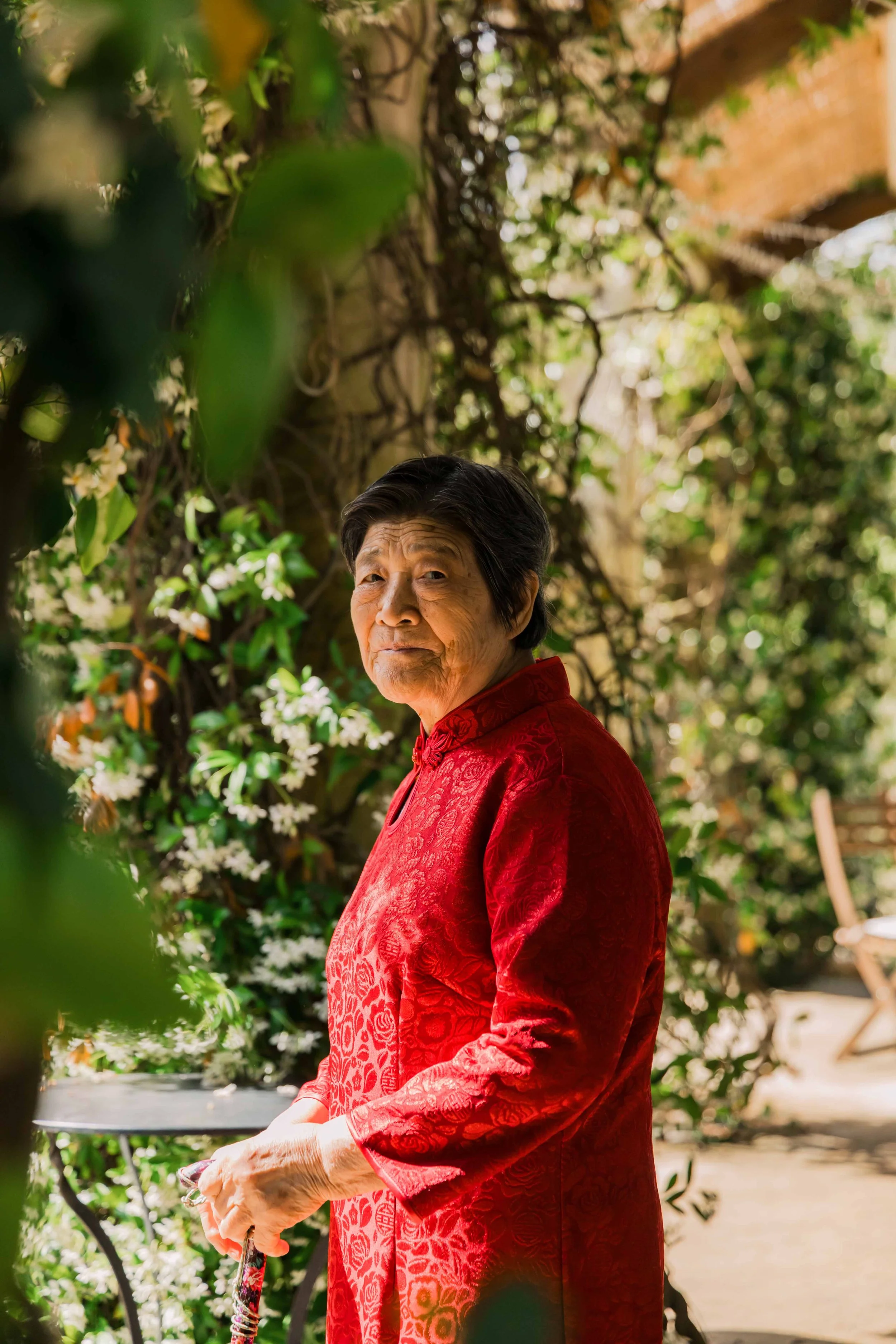 An elderly woman wearing a red traditional dress stands outdoors in a garden with green foliage and white flowers, looking at the camera.