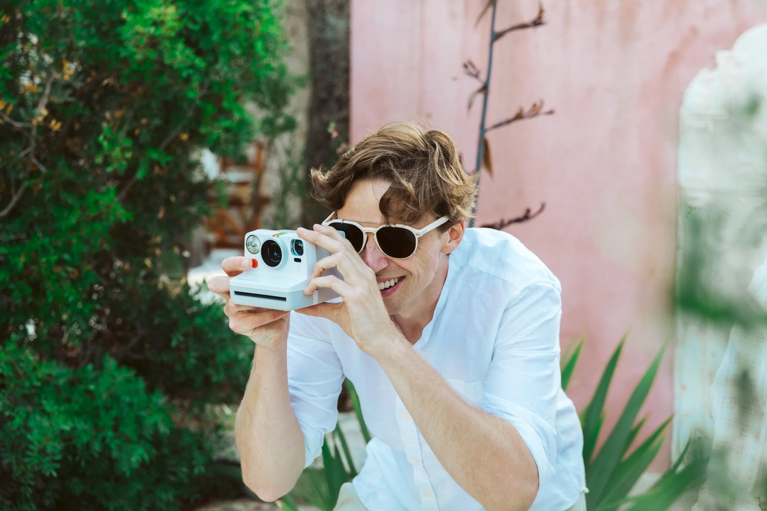 A young man wearing sunglasses smiling and taking a photo outdoors with a vintage instant camera.