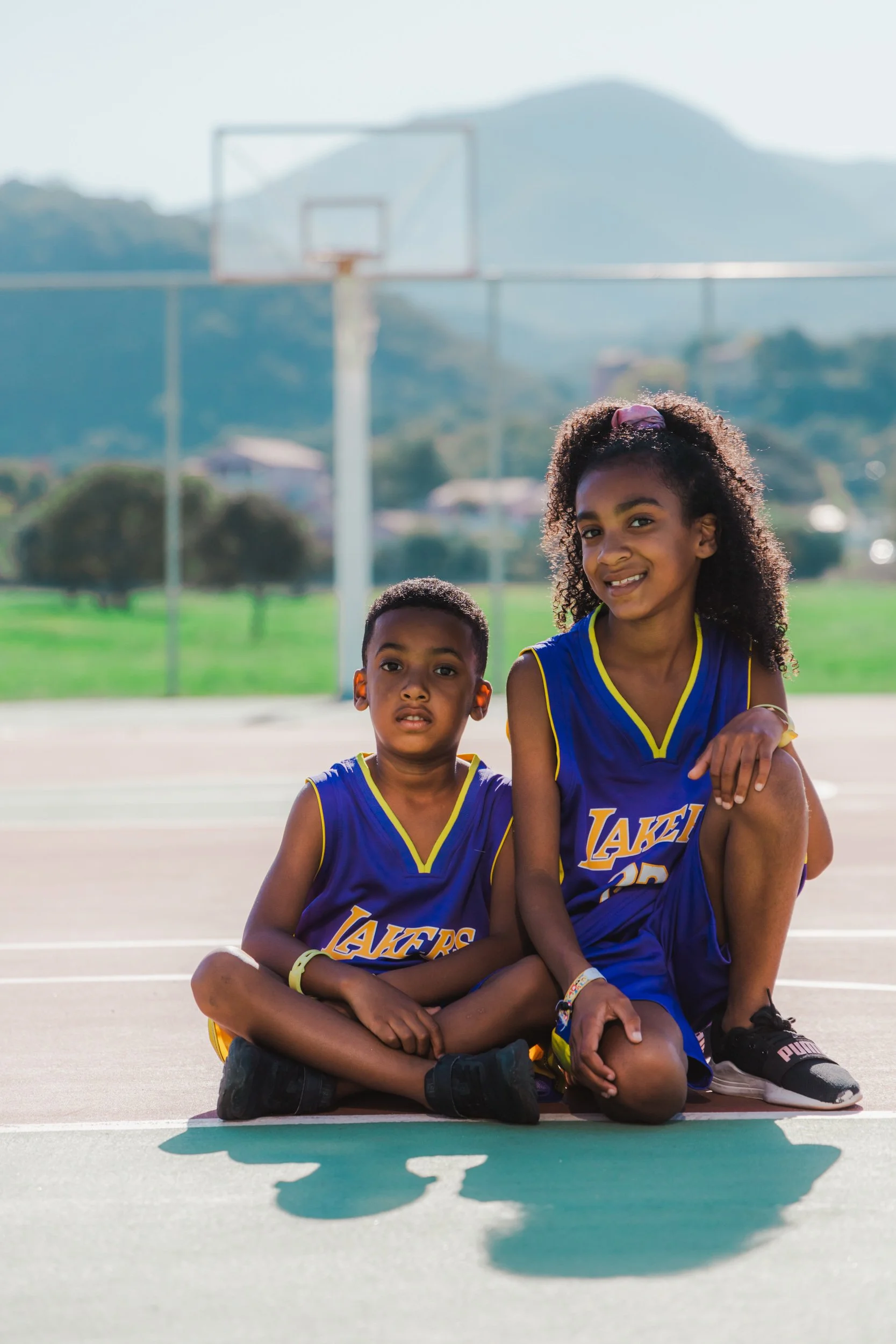 Two children sitting on a basketball court, wearing Los Angeles Lakers jerseys, with a basketball hoop and mountains in the background.