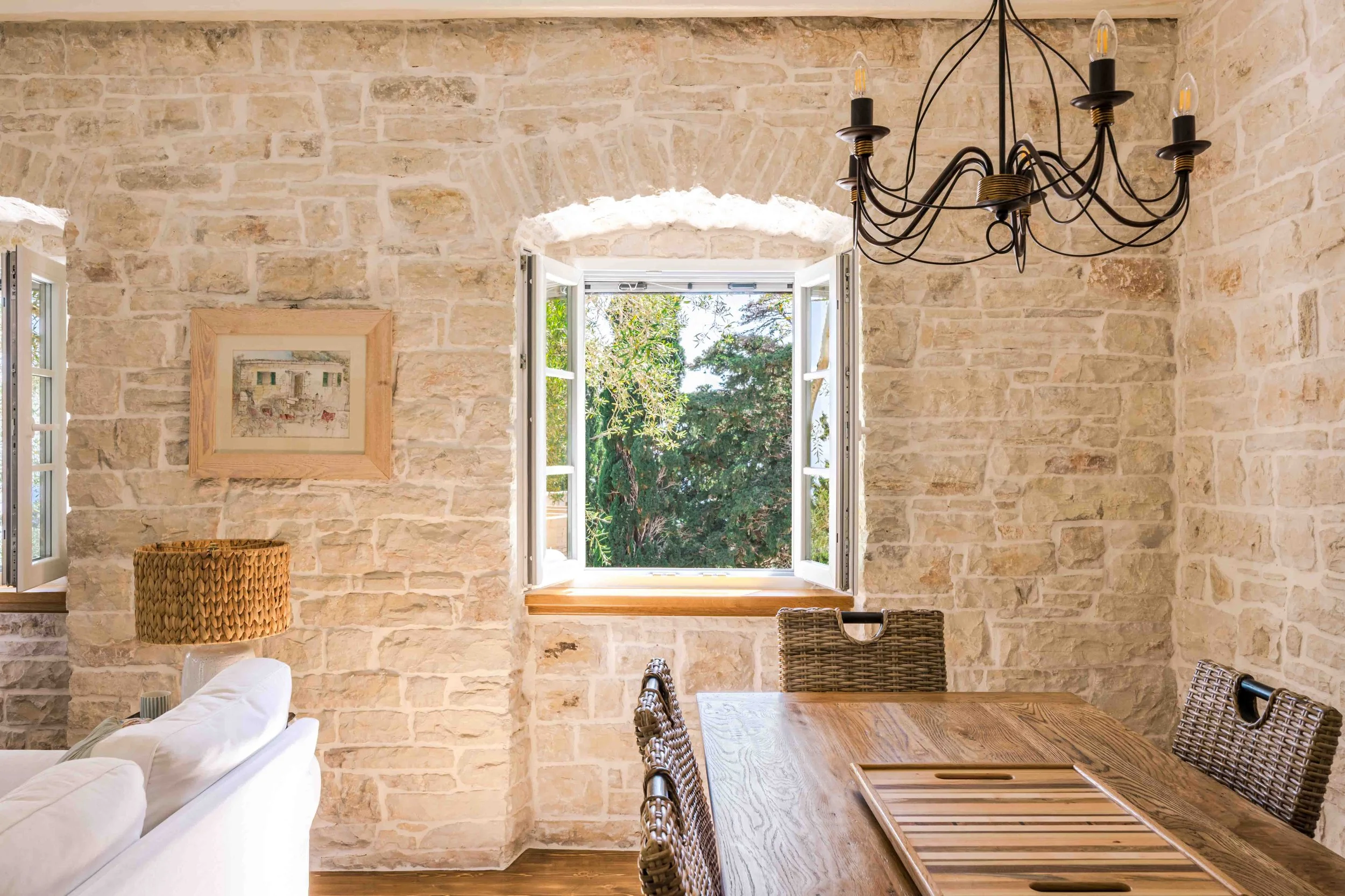 A dining room with exposed light-colored brick walls, an open window with a view of trees, a black wrought iron chandelier, a wooden dining table with woven chairs, and a white sofa with a wicker lampshade nearby.
