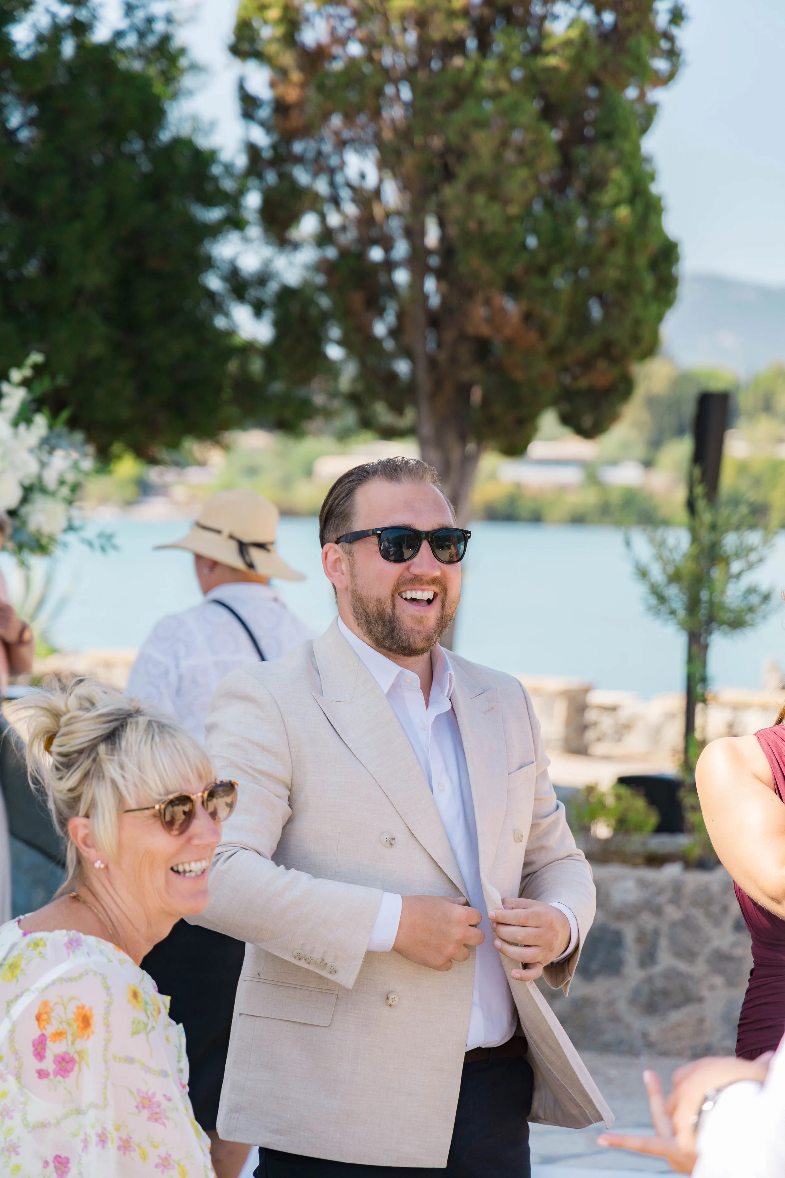 A man in a light-colored suit and sunglasses smiling and talking at an outdoor event near a body of water. A woman with blonde hair and sunglasses is smiling nearby, wearing a white dress with floral patterns.