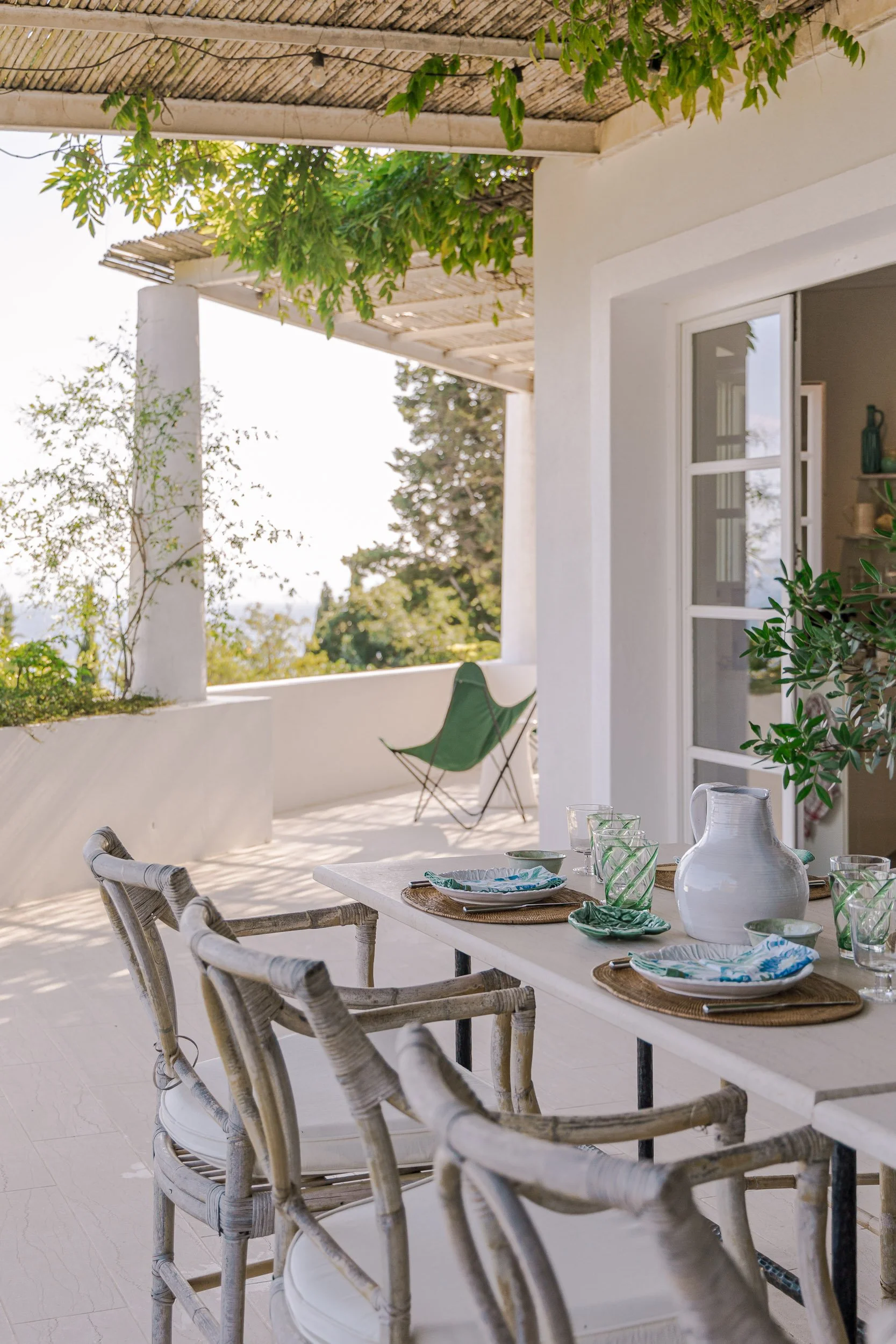 A table set for a meal on a balcony, with chairs, plates, glasses, and a pitcher, overlooking a sunny outdoor space with greenery and a lounge chair.