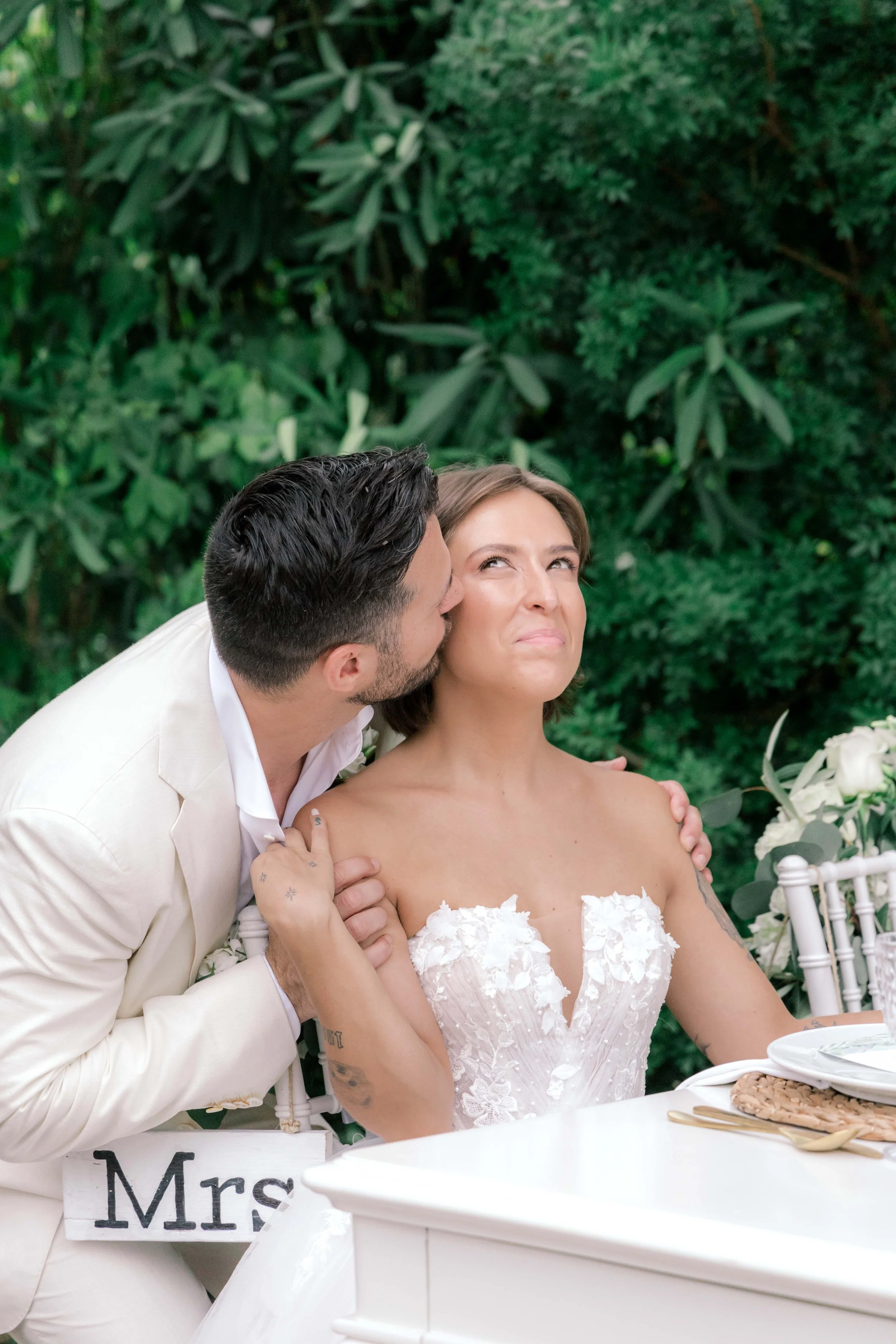A man kissing a woman on the cheek at a wedding reception outdoors, with greenery in the background. The woman is seated at a white table with flowers and a "Mrs" sign, wearing a white wedding dress.