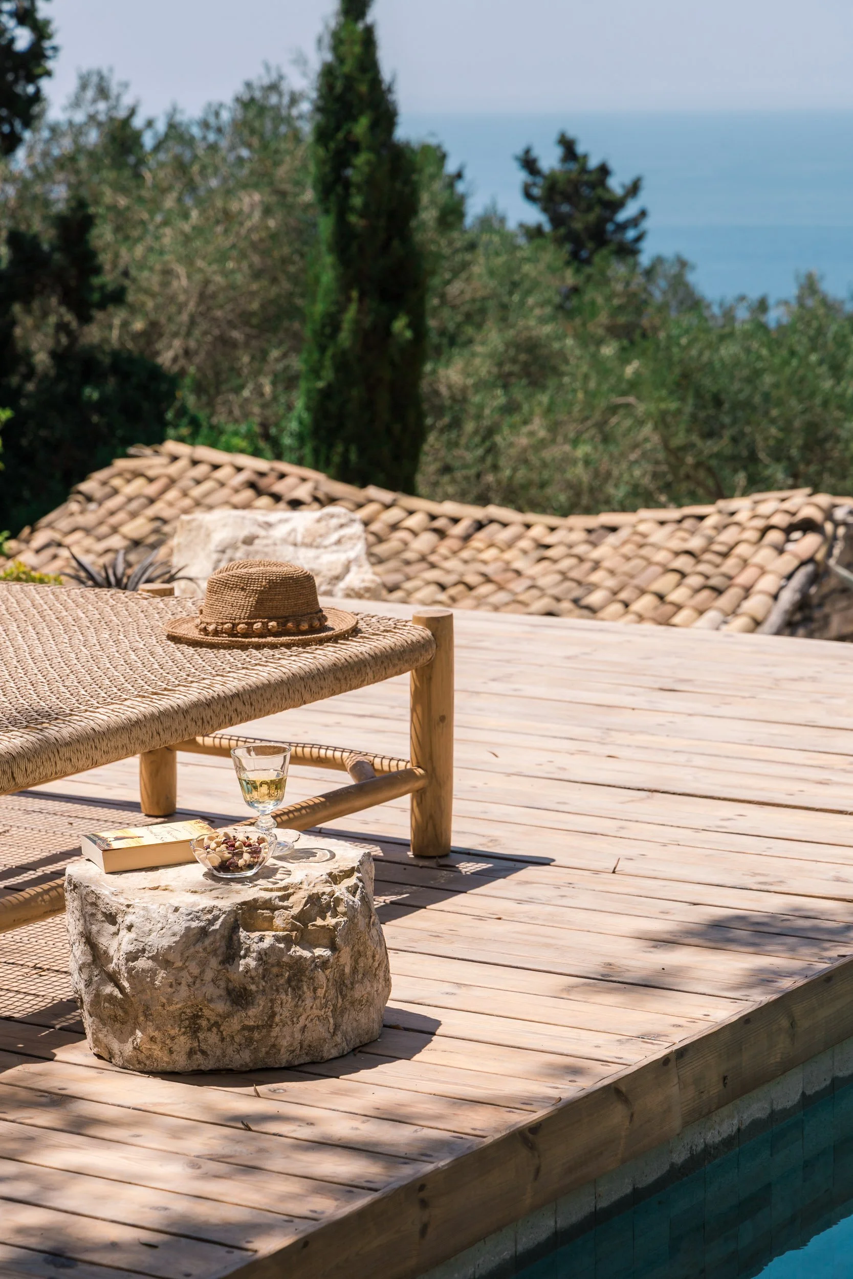 A wooden poolside deck with a woven bench, a straw hat, a glass of white wine, a bowl of snacks, and a book, overlooking trees and the ocean in the background.