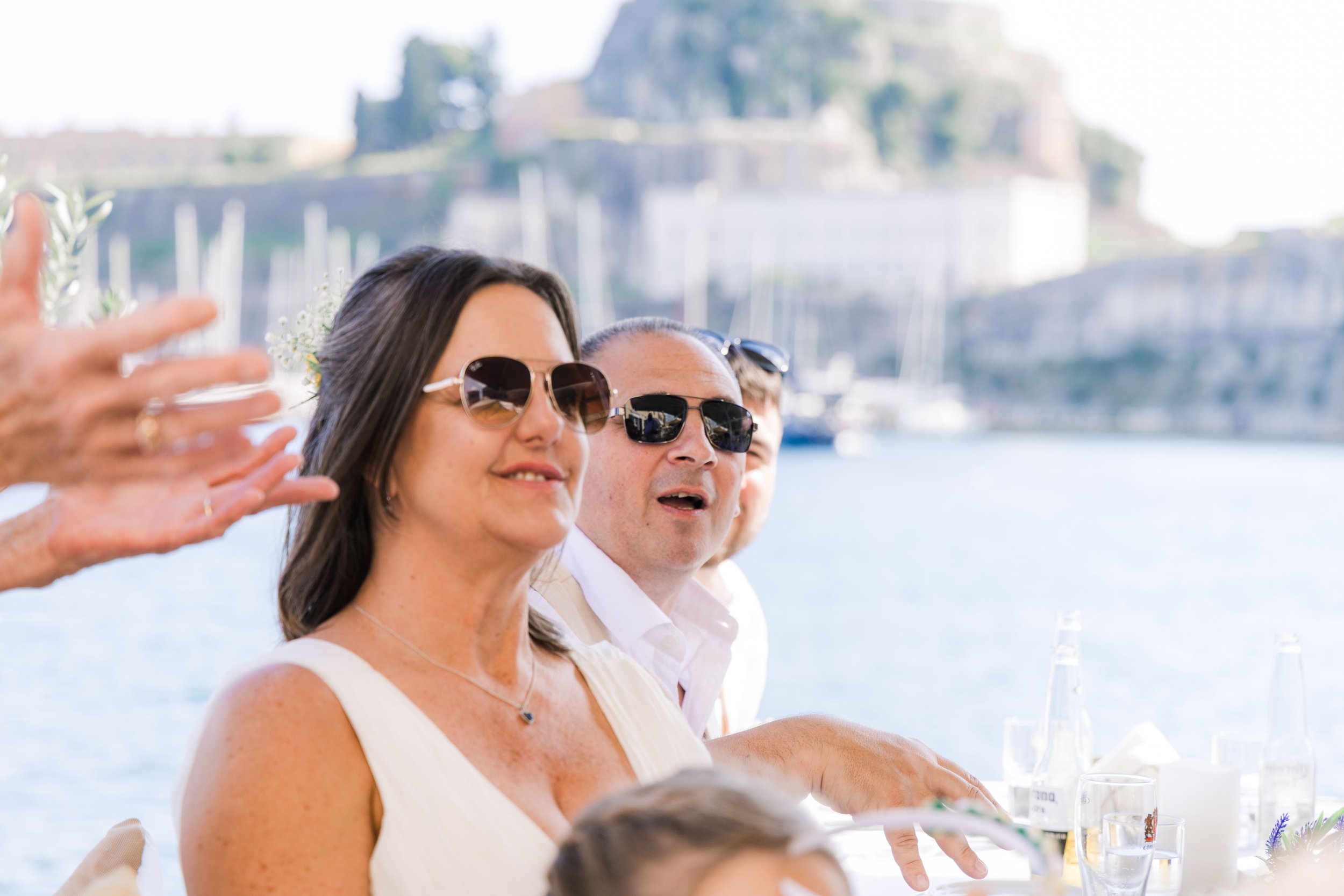 Group of people enjoying an outdoor gathering by the water with boats and hilly landscape in the background, some wearing sunglasses.