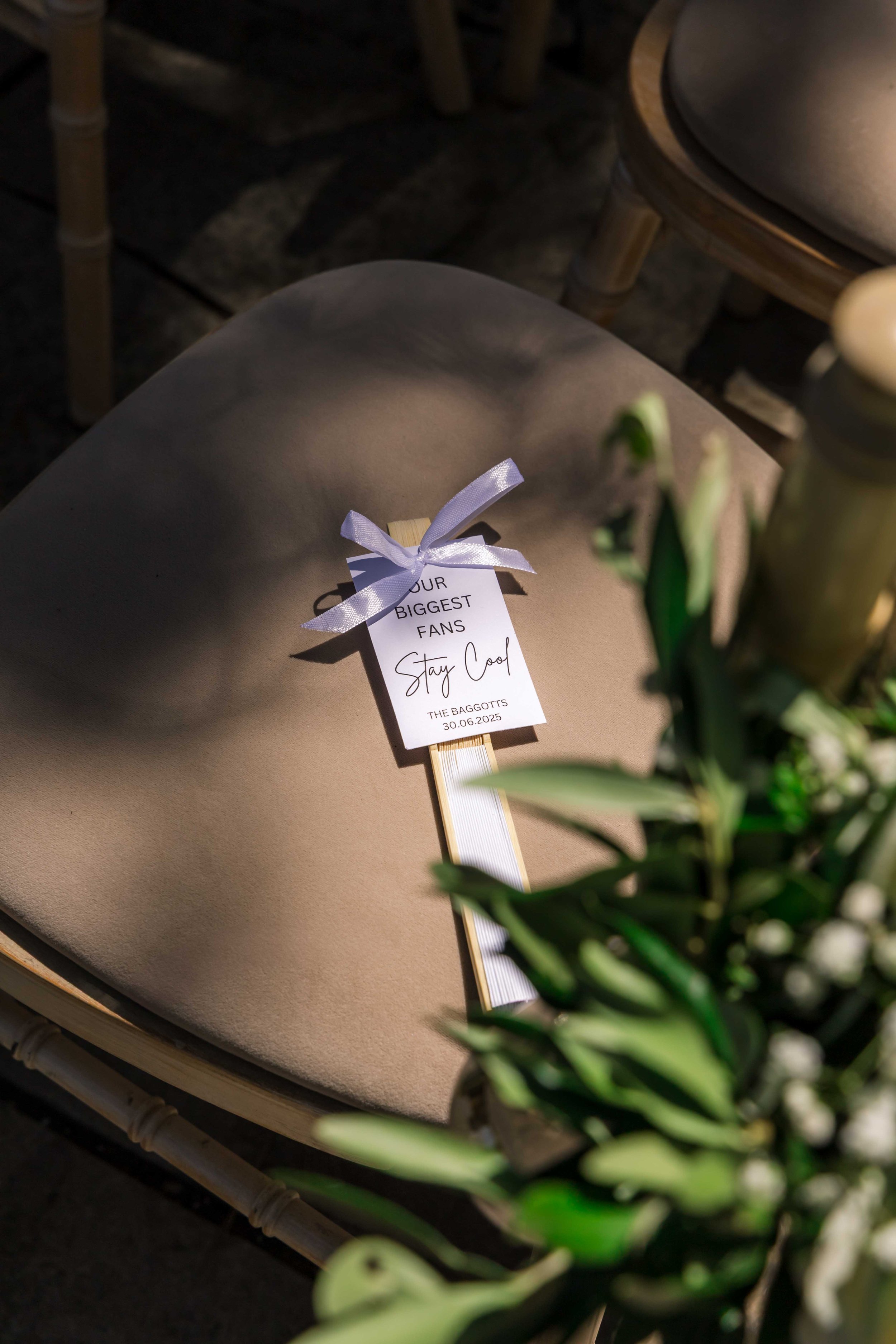 A beige cushioned chair with a small wooden sign tied with a lavender ribbon, reading "Your Biggest Fans Stay Cool" and dated June 30, 2025, placed on it. A blurred green plant is in the foreground.