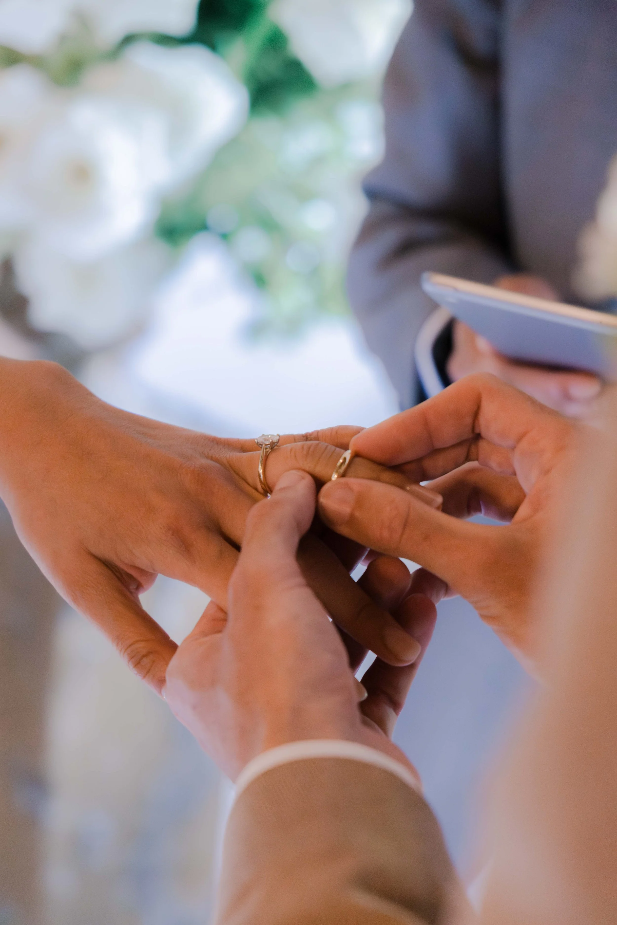 A close-up of a wedding ceremony with one person placing a ring on another person's finger. The background shows blurry white flowers and greenery.