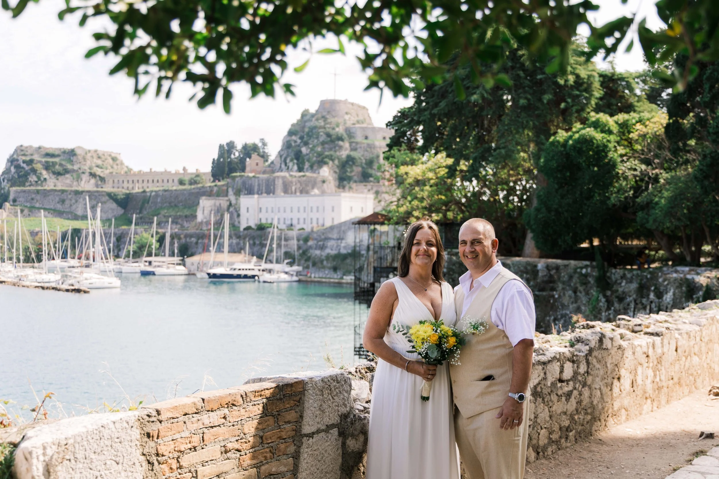 A couple dressed in wedding attire standing next to each other on a waterfront, smiling, with a castle and boats in the background, surrounded by greenery.
