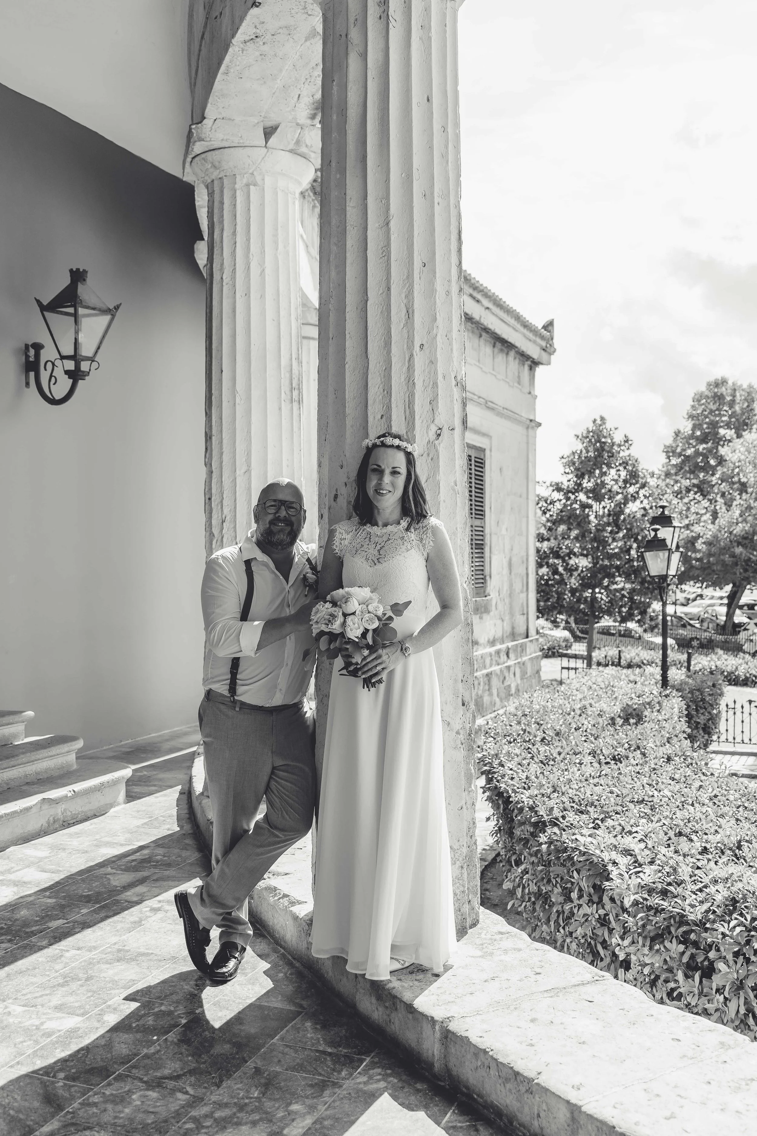 A couple, possibly at a wedding, standing outside near a classical building. The woman is in a white dress with lace, holding a bouquet and wearing a floral crown. The man is in a shirt with suspenders, leaning against a column, smiling.