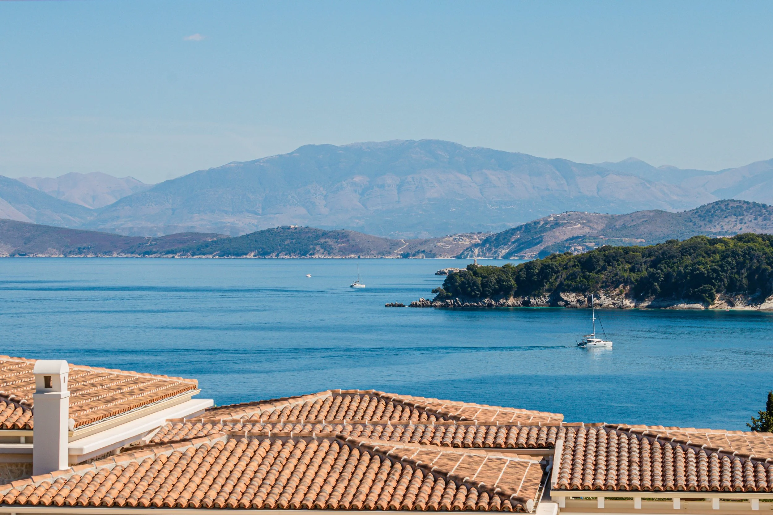 View of a lake or bay with sailboats, lush islands, and distant mountains, seen from a balcony with tiled roof and chimney in the foreground.