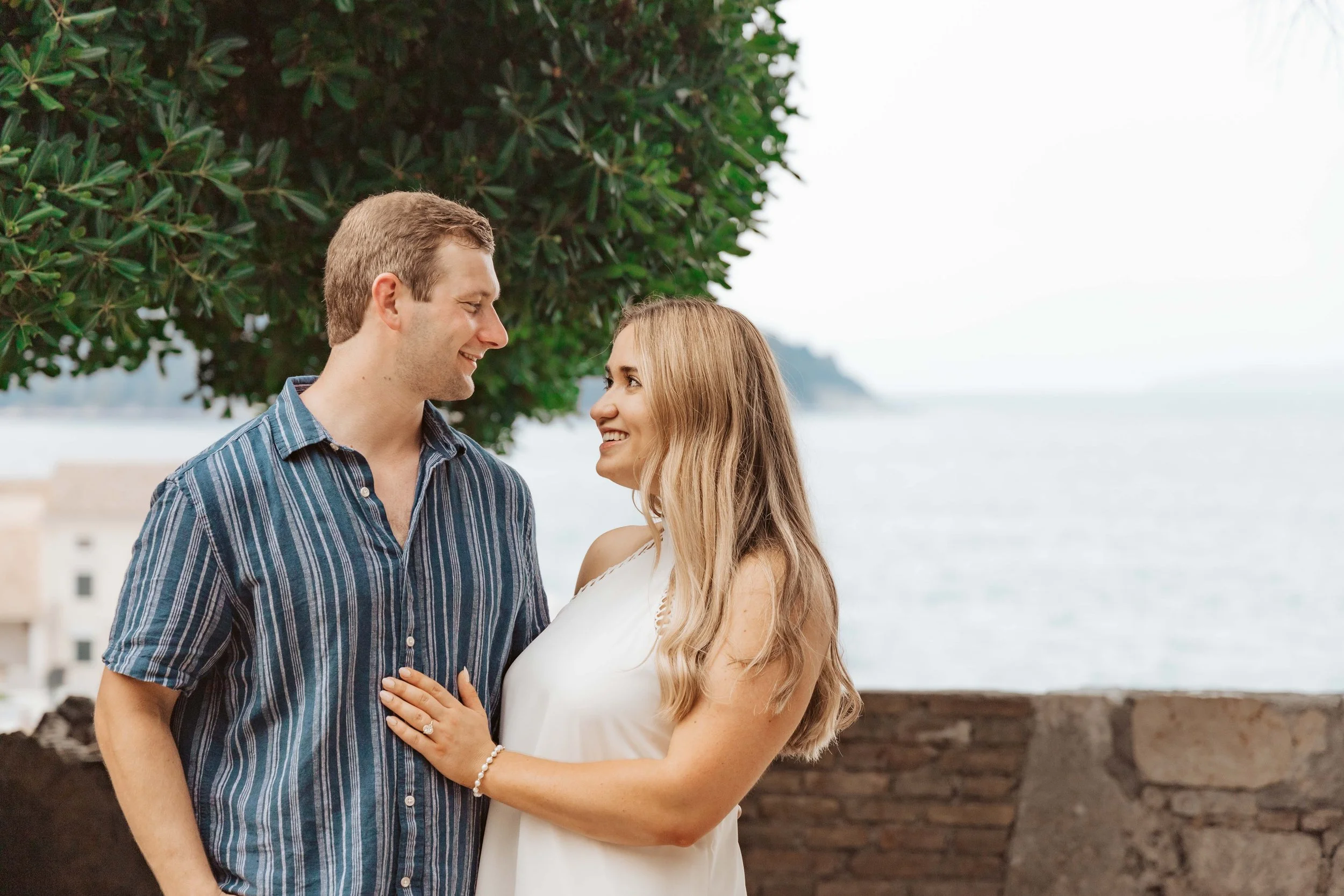 A couple stands outdoors near the ocean, smiling and gazing into each other's eyes. The man has short brown hair, wears a blue striped shirt, and the woman has long blonde hair, wears a white dress, and a pearl bracelet. They are standing close by a 