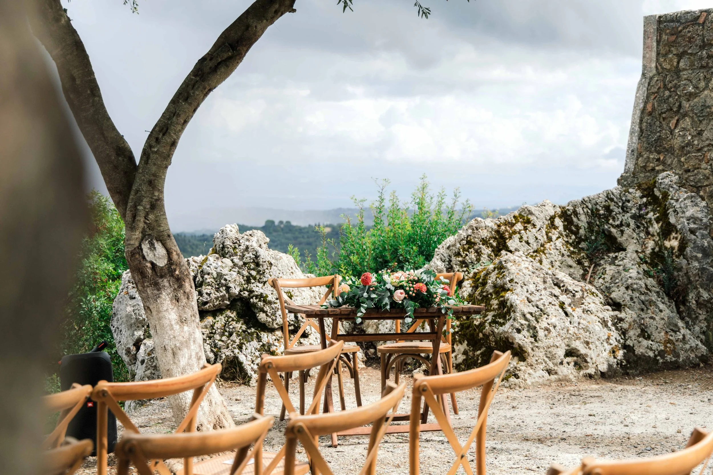 An outdoor wedding setup with a wooden table decorated with flowers and greenery, surrounded by wooden chairs, set against rocky terrain and lush greenery under a cloudy sky.