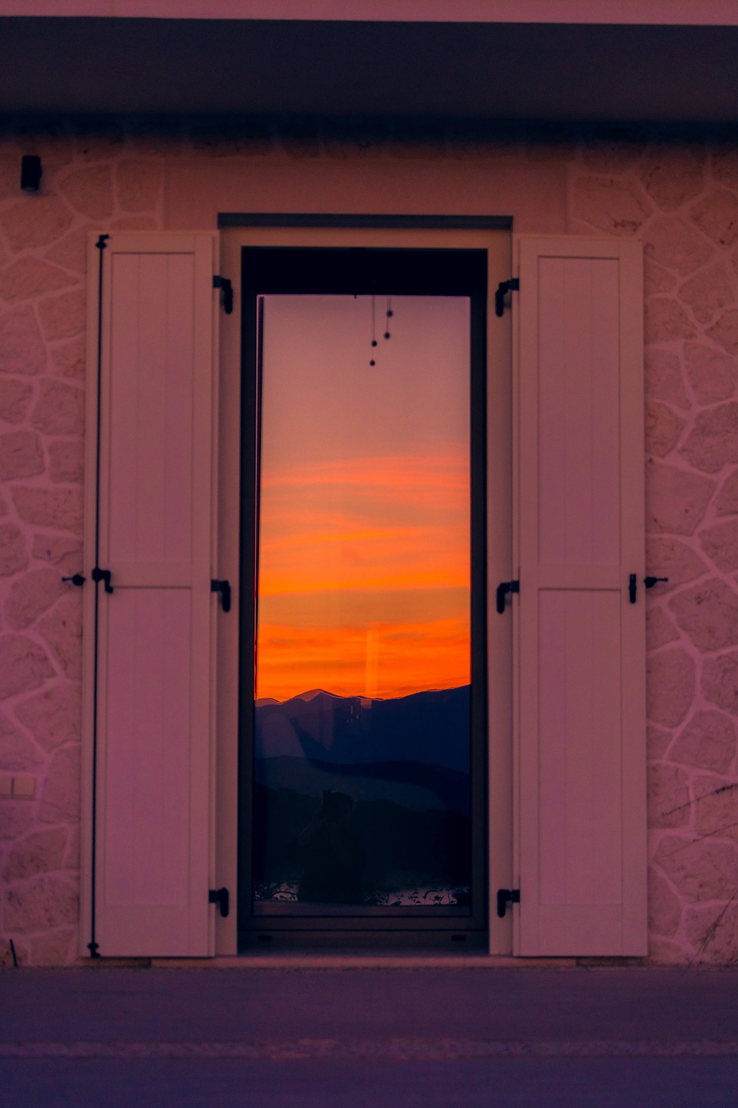 Sunset view seen through a glass door with white shutters, featuring a vibrant sky with orange, pink, and purple hues over silhouette mountains.