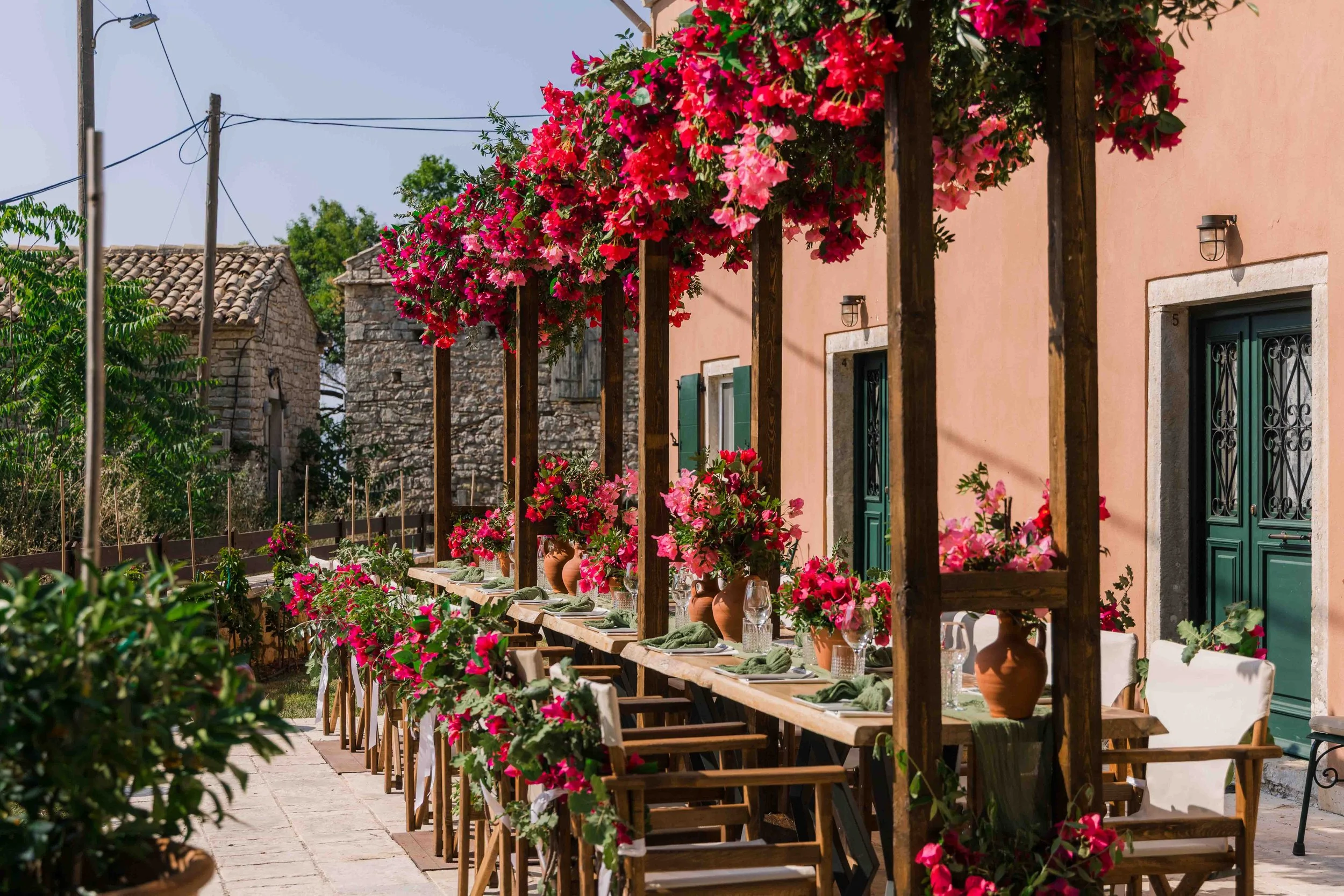 Outdoor dining setup with a long wooden table, chairs, and vases of pink and red flowers. The table is under a pergola decorated with pink flowering plants, adjacent to pink-colored building walls with green doors. The scene is sunlit with some old s