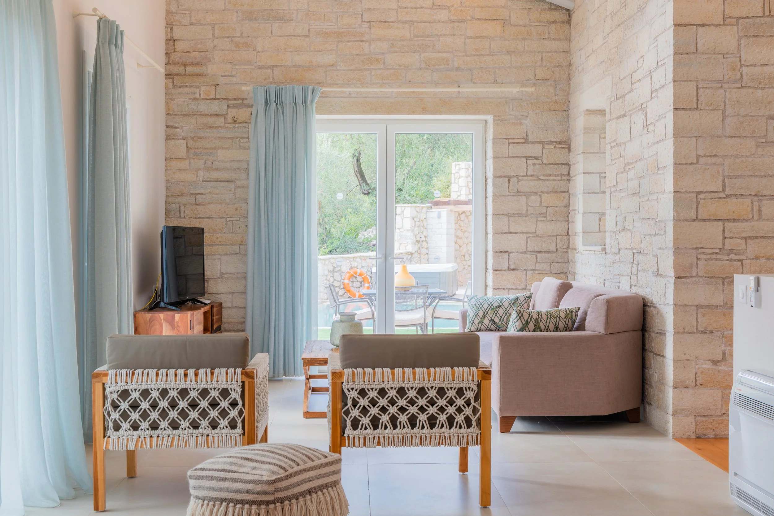Living room with stone walls, a sliding glass door leading to an outdoor patio with a table and chairs, a beige sofa with decorative pillows, two woven armchairs, a small wooden side table, a TV on a wooden console, light blue curtains, and light-col
