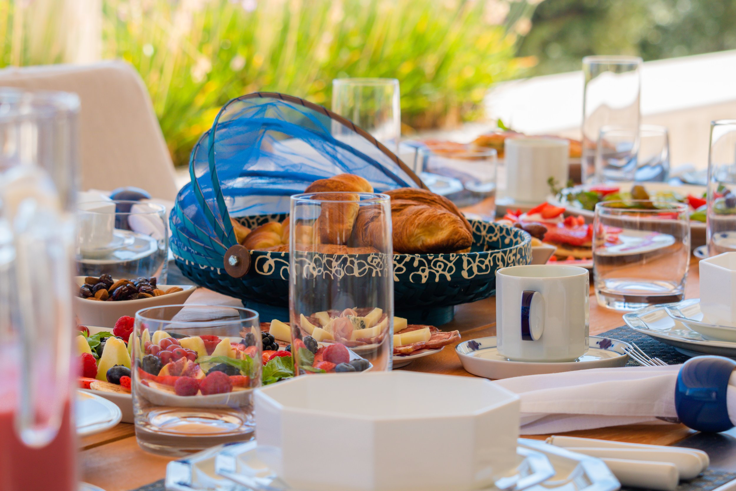 Table set for breakfast with assorted pastries, fresh fruit, coffee mug, water glasses, and plates with salads, outdoors with greenery in the background.