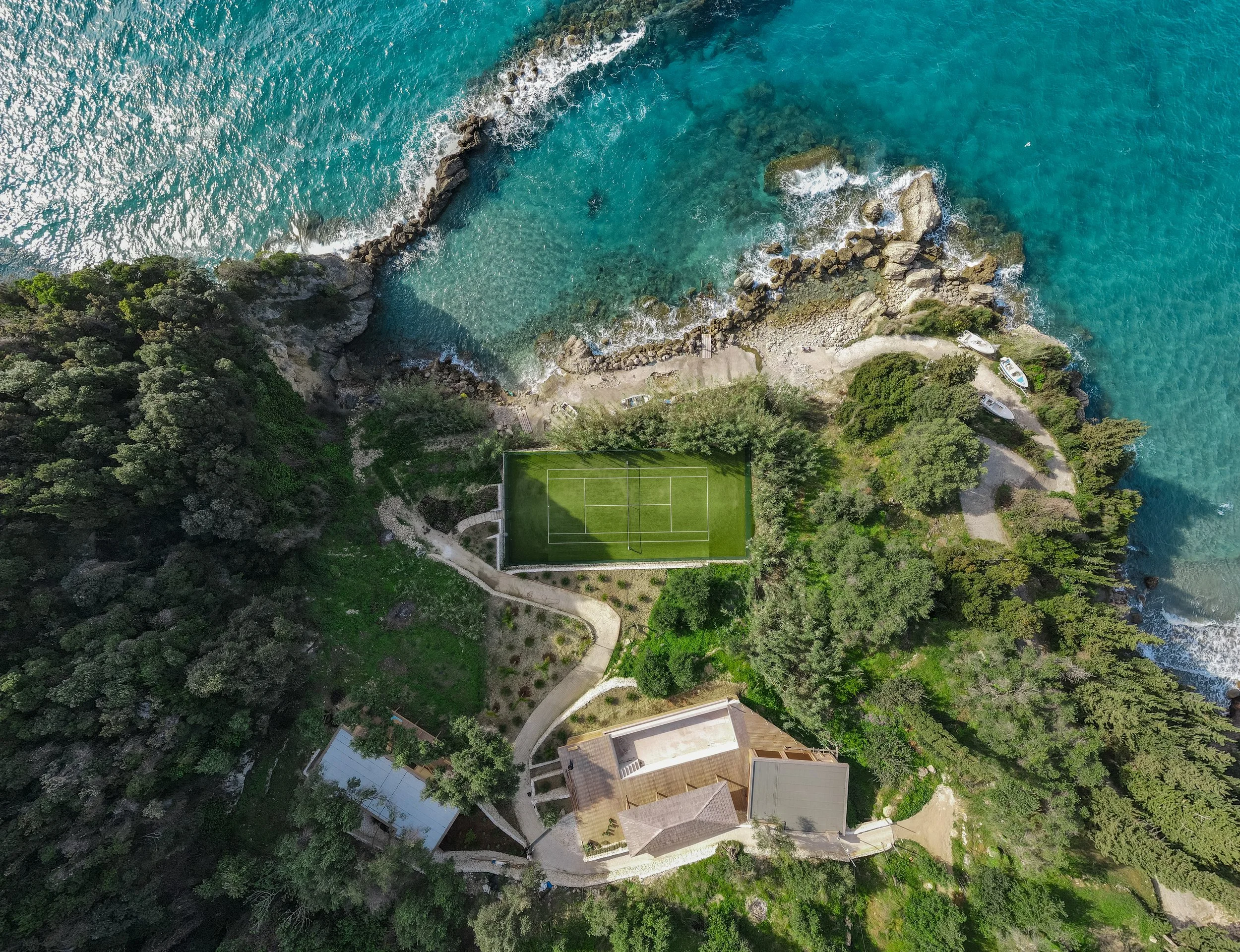 Aerial view of a coastal property featuring a tennis court, surrounded by trees and ocean waves crashing against rocky shoreline.