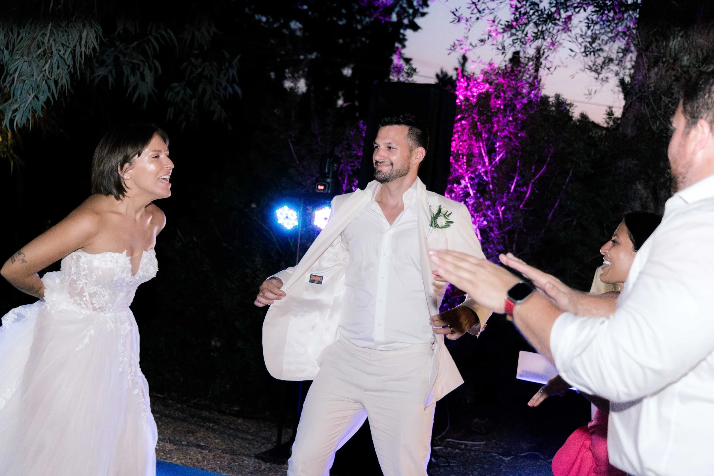 People dancing and celebrating at a wedding reception outdoors during evening, with colorful purple and blue lighting and trees in the background.