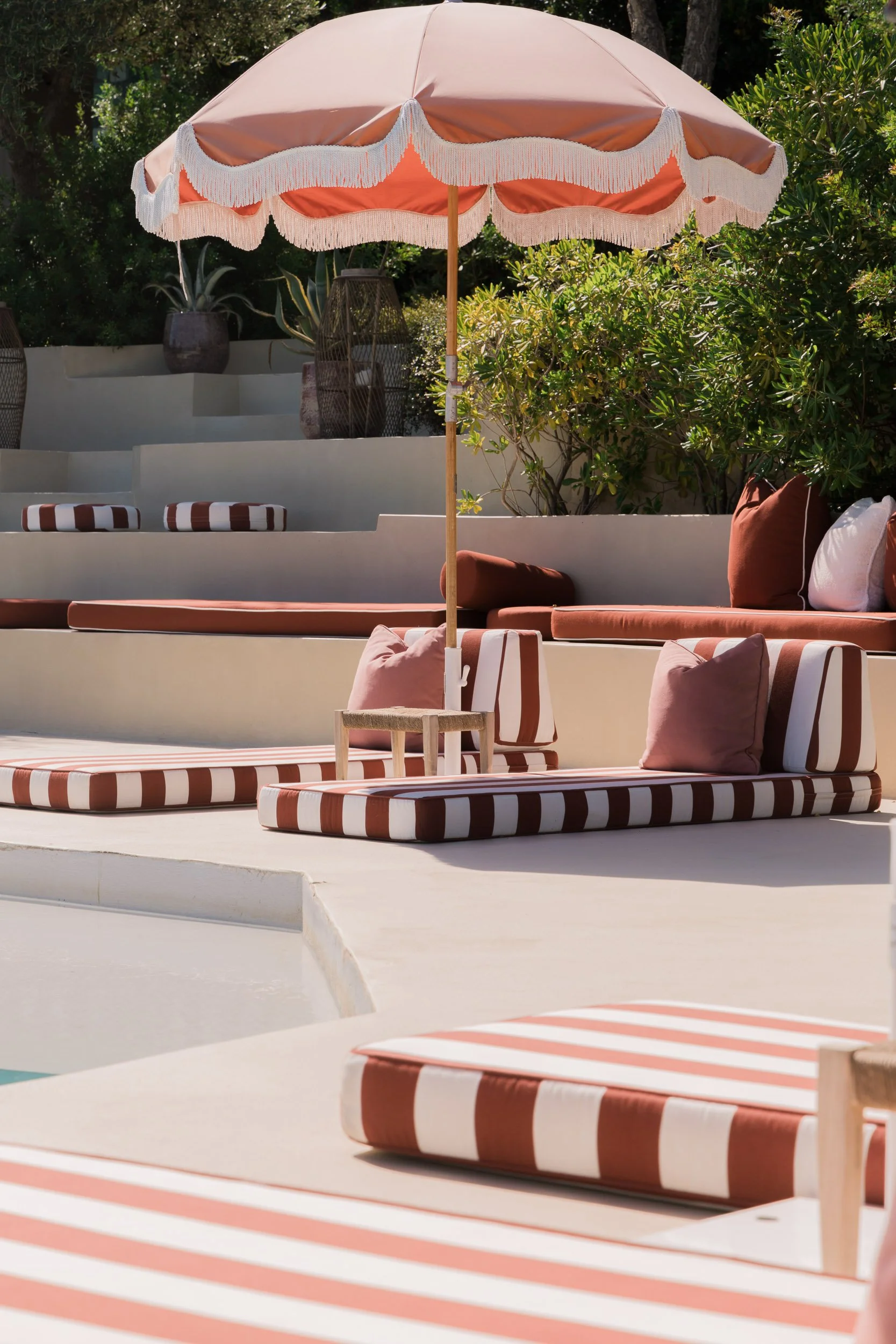 Poolside area with striped cushioned loungers, pink pillows, a large pink and white umbrella, and lush green plants.