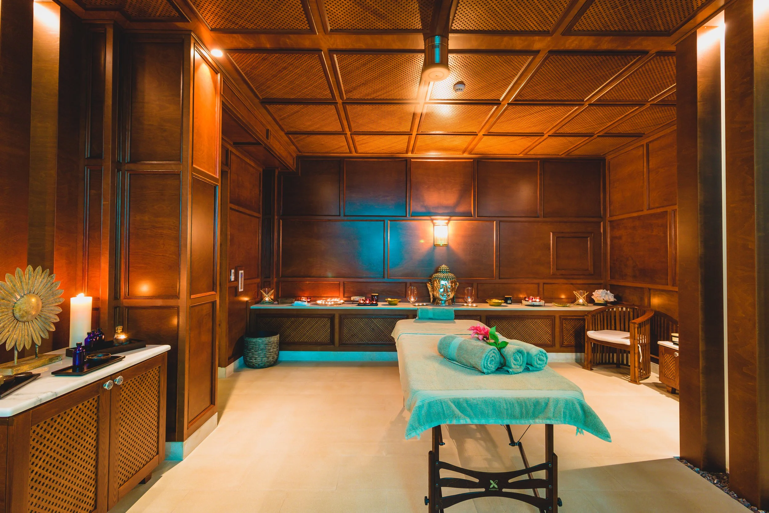 A luxurious spa room with a massage table, rolled towels, and decorative elements with warm lighting and wooden paneling on the walls.