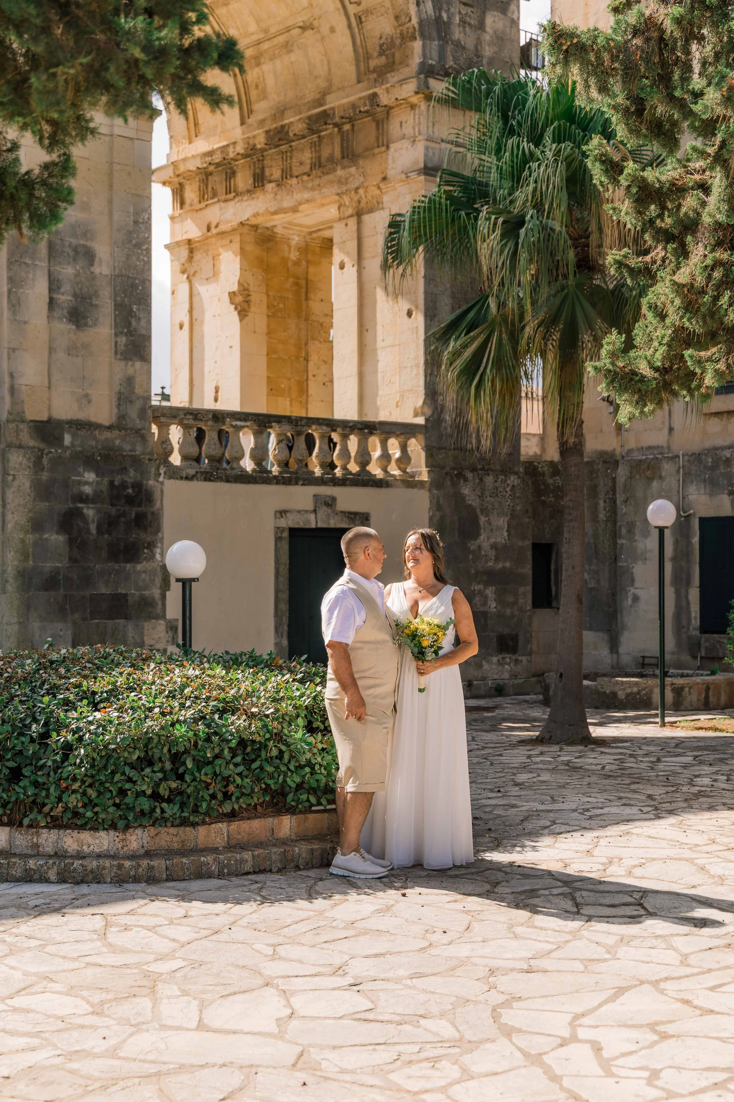 A couple dressed in wedding attire standing outside in front of historic stone architecture, with the woman holding a bouquet and gazing at the man.