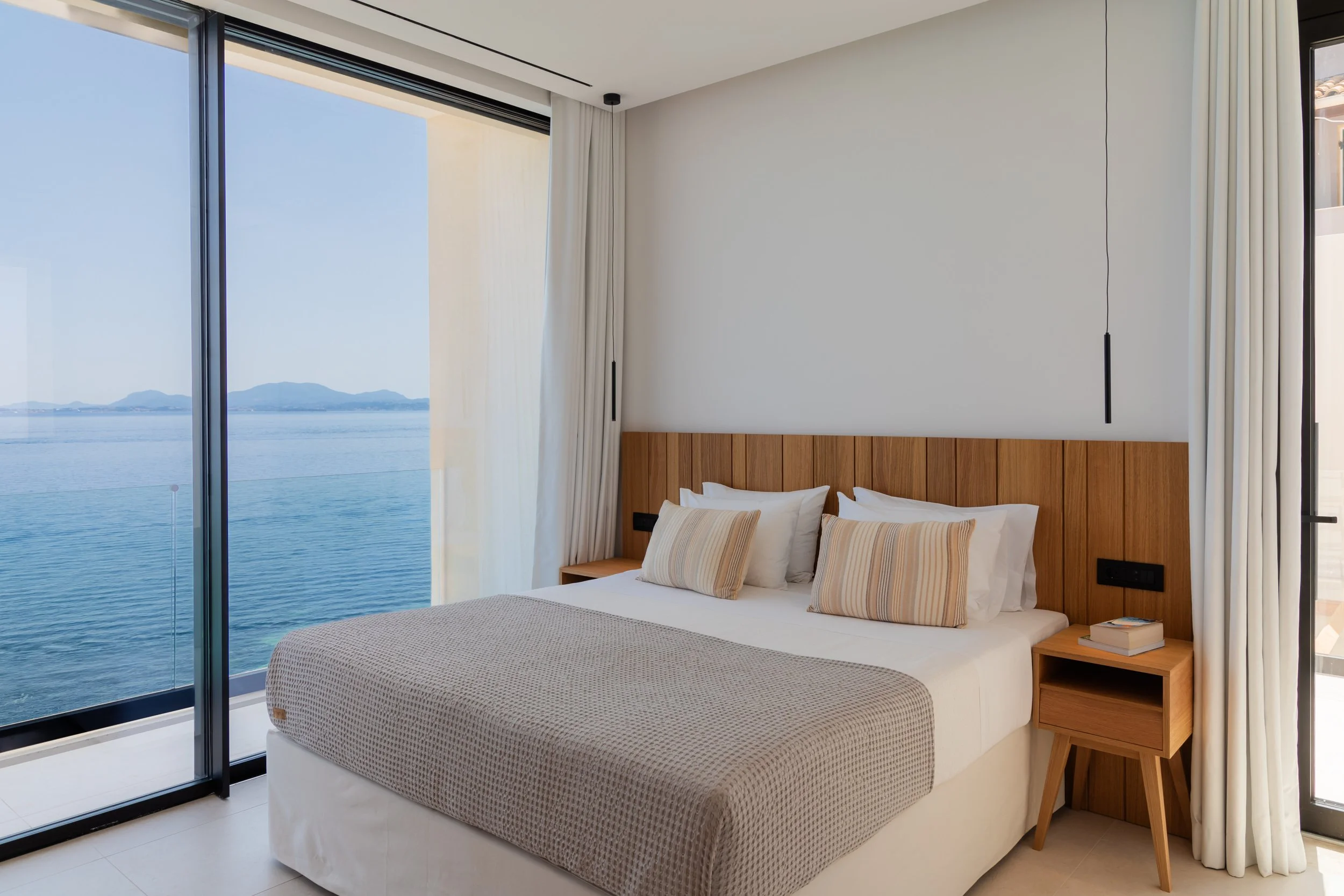 A bedroom with a large bed, beige and white striped pillows, and a view of a body of water and mountains through a sliding glass door.