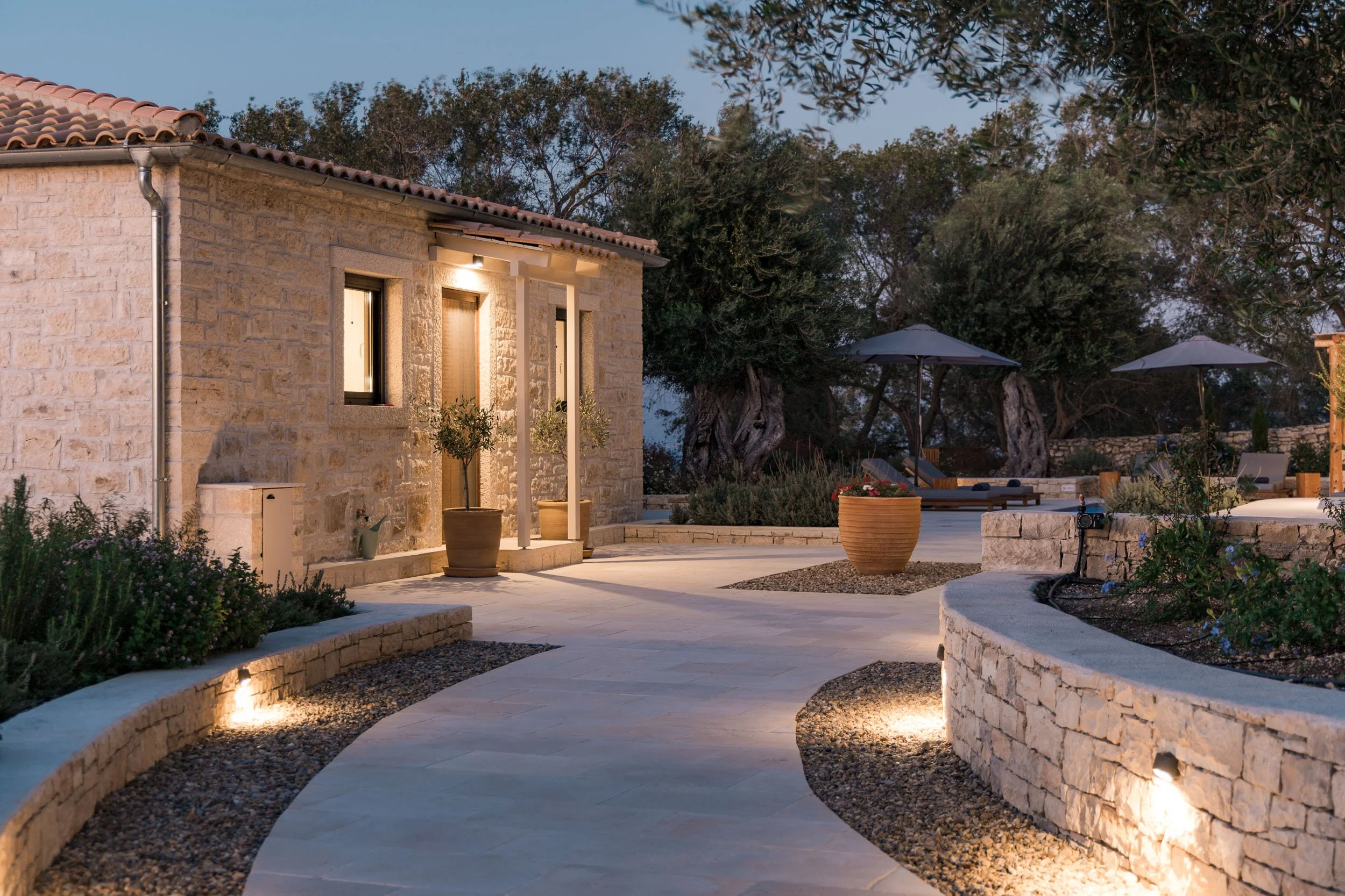 A stone house with warm lights on a patio at dusk, surrounded by trees, with potted plants, outdoor lounge chairs, and umbrellas.
