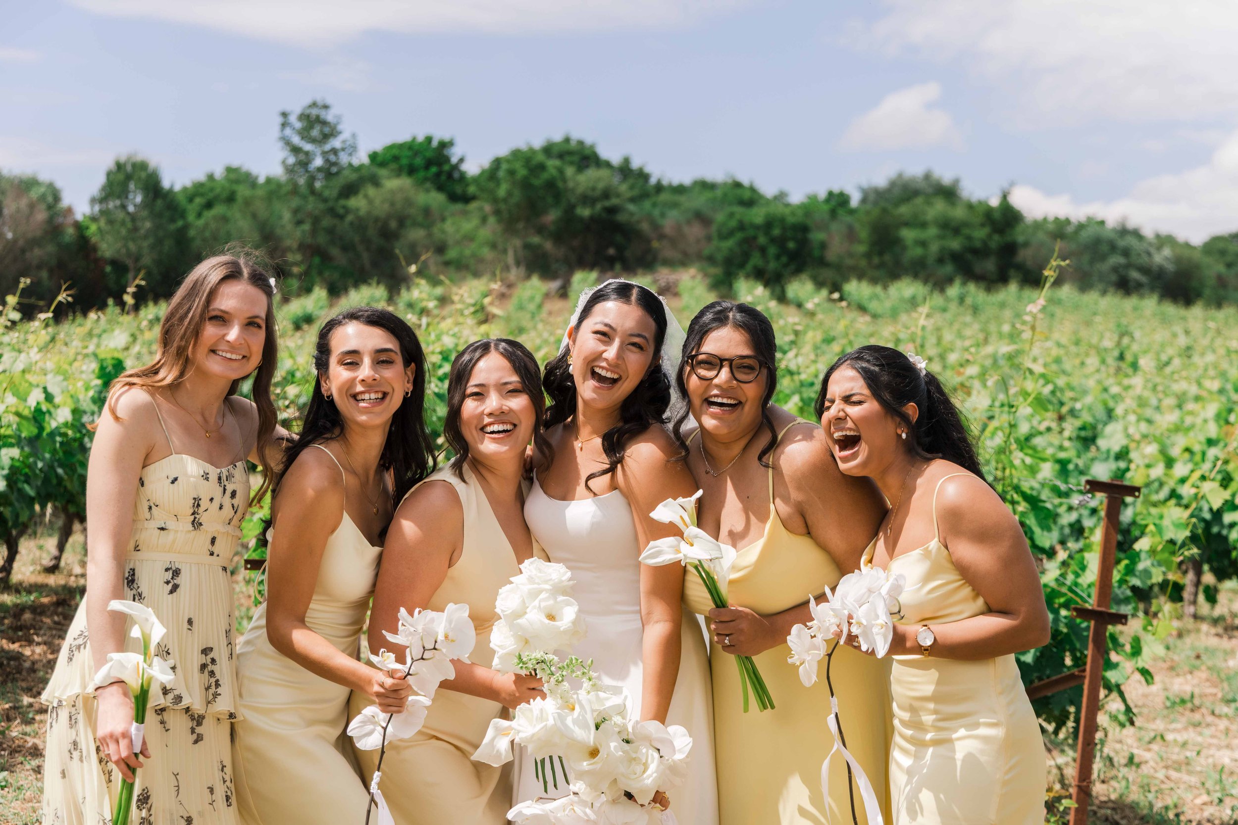 A group of six women dressed in light-colored dresses standing in a vineyard, smiling and laughing during a wedding celebration, holding white flowers, with green vines and a partly cloudy sky in the background.