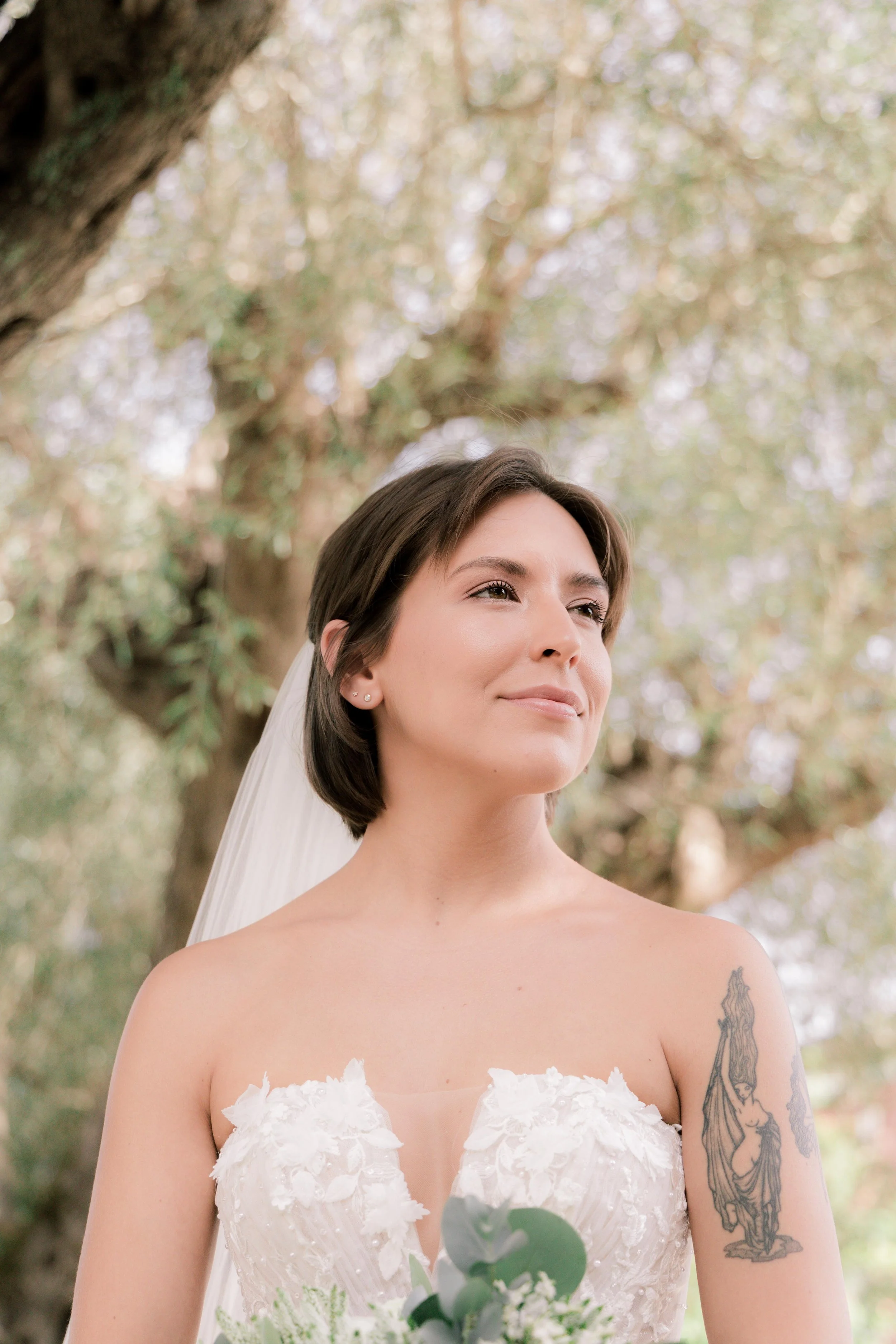 A woman in a white wedding dress with floral embellishments, short dark hair, and a tattoo on her upper arm, standing outdoors with trees and foliage in the background.