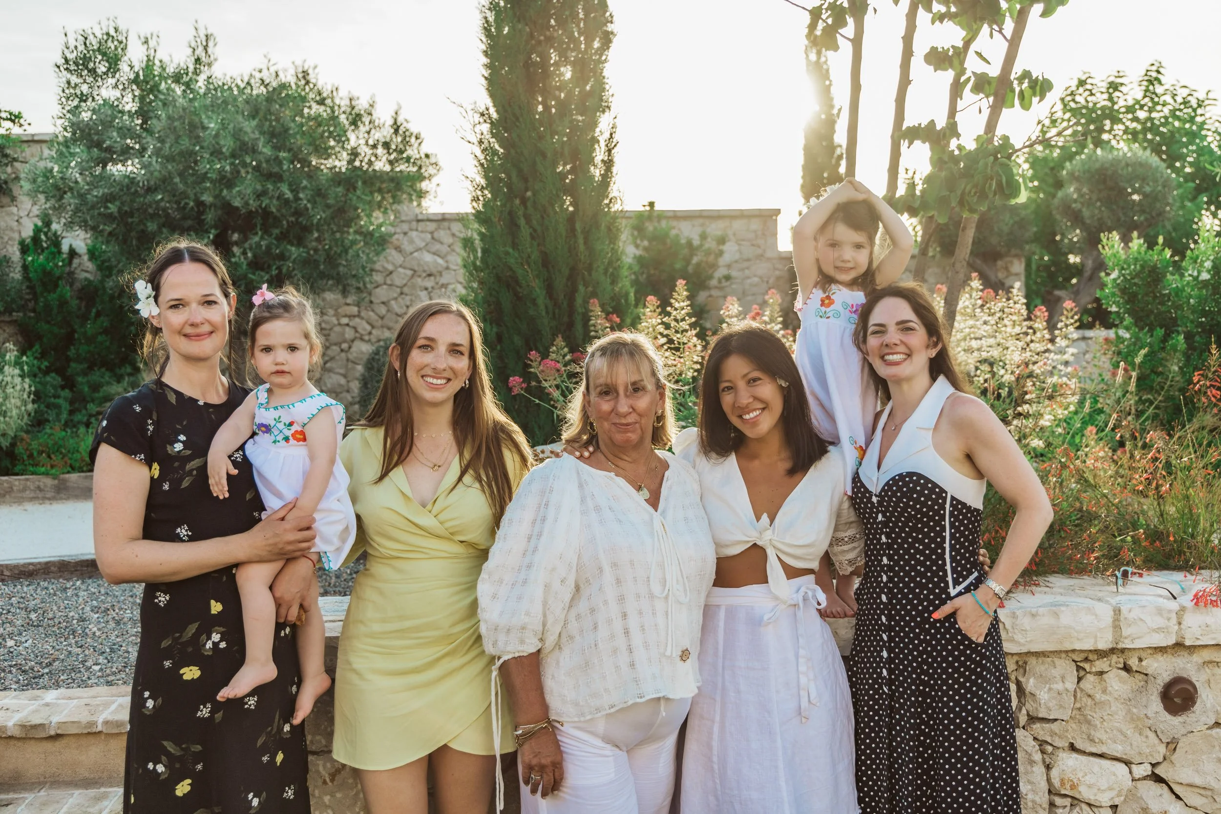 Group of seven women and two young girls standing outdoors in a garden with greenery and stone wall, smiling in sunlight.