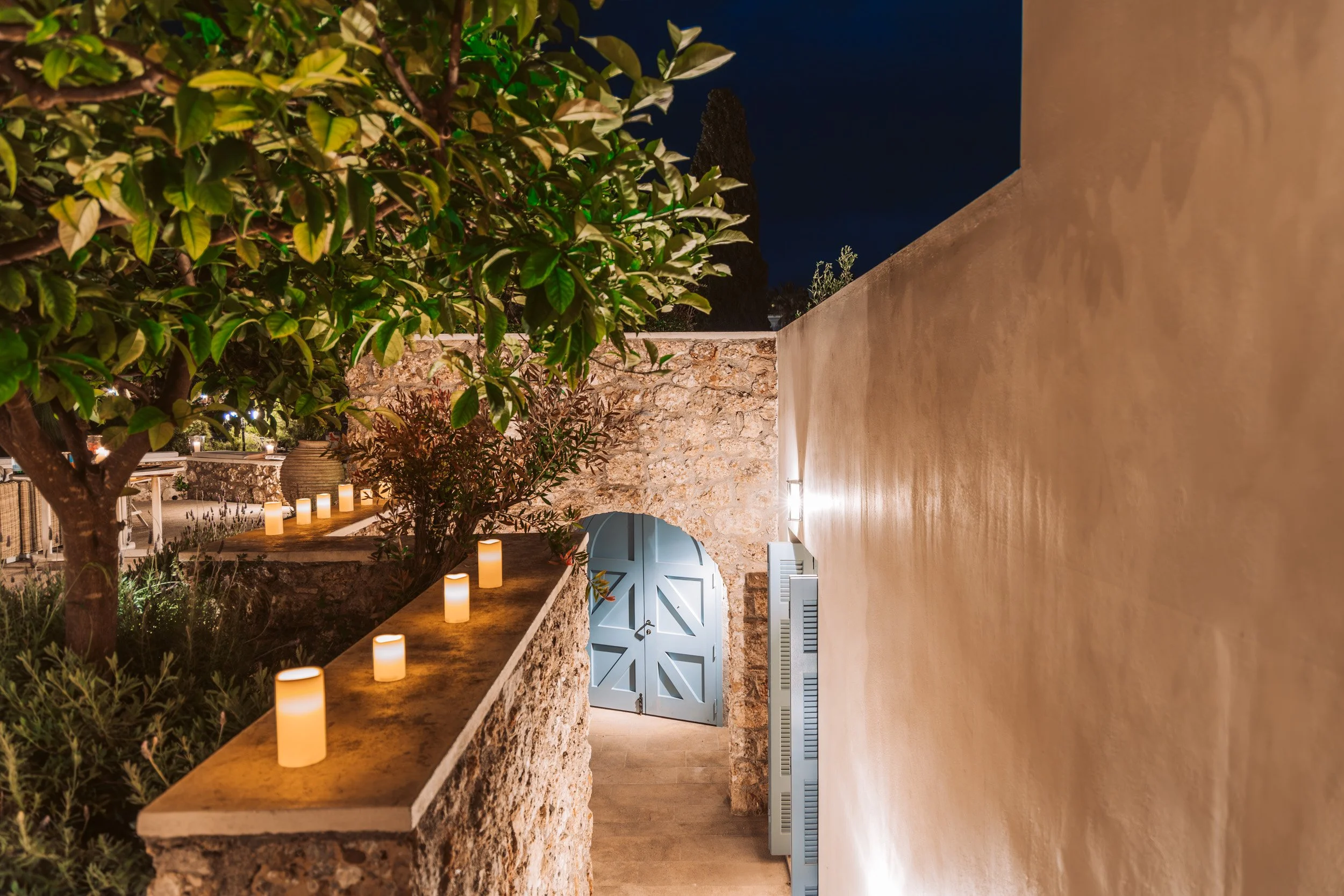 Night view of a terrace with a stone wall, a blue wooden door, potted plants, candle lights on a stone ledge, and a large leafy tree.