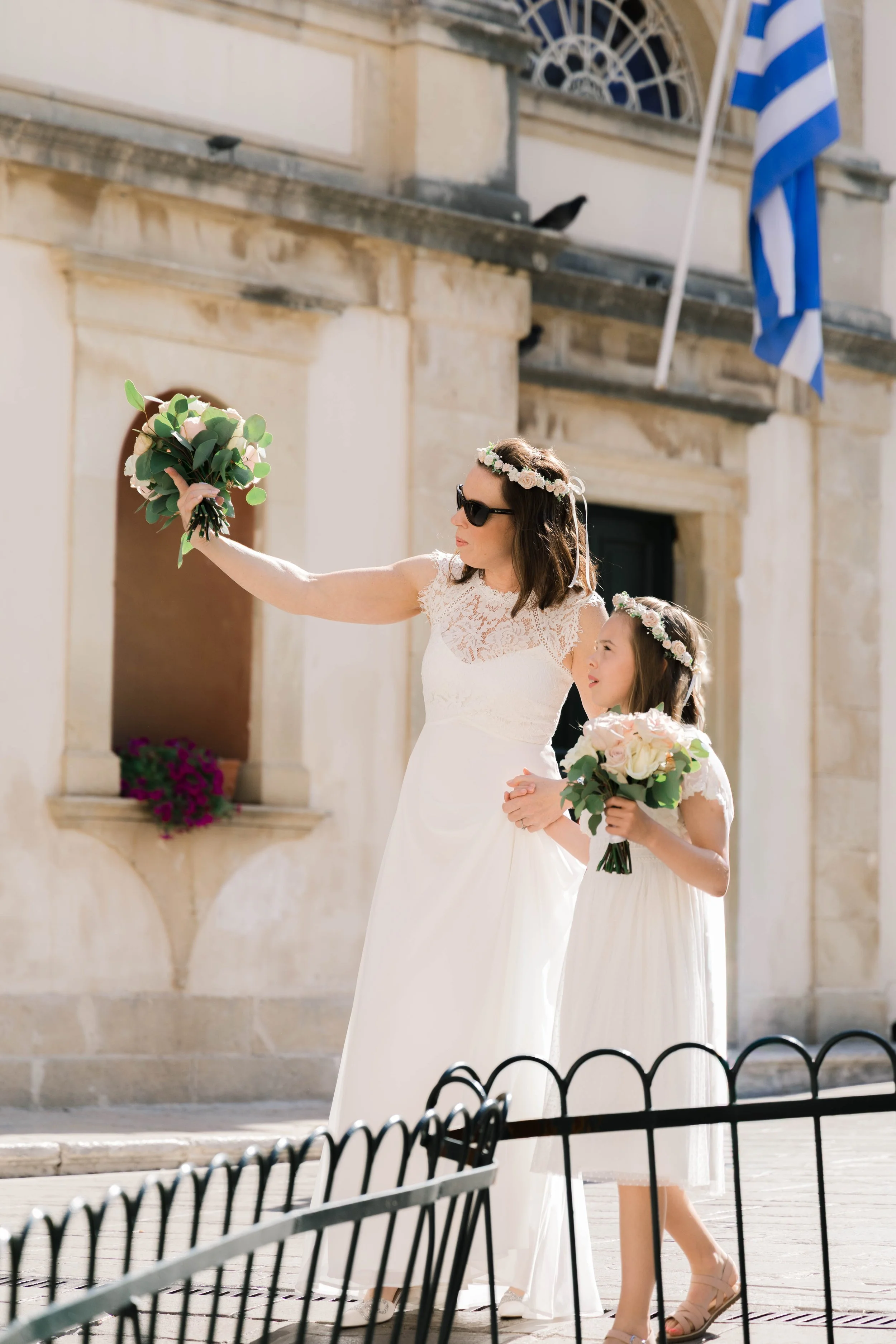 A woman in a white lace dress and sunglasses holding a bouquet, and a young girl in a white dress with a floral crown and holding a bouquet, standing in front of an ornate building with Greek flag and a standing black fence.
