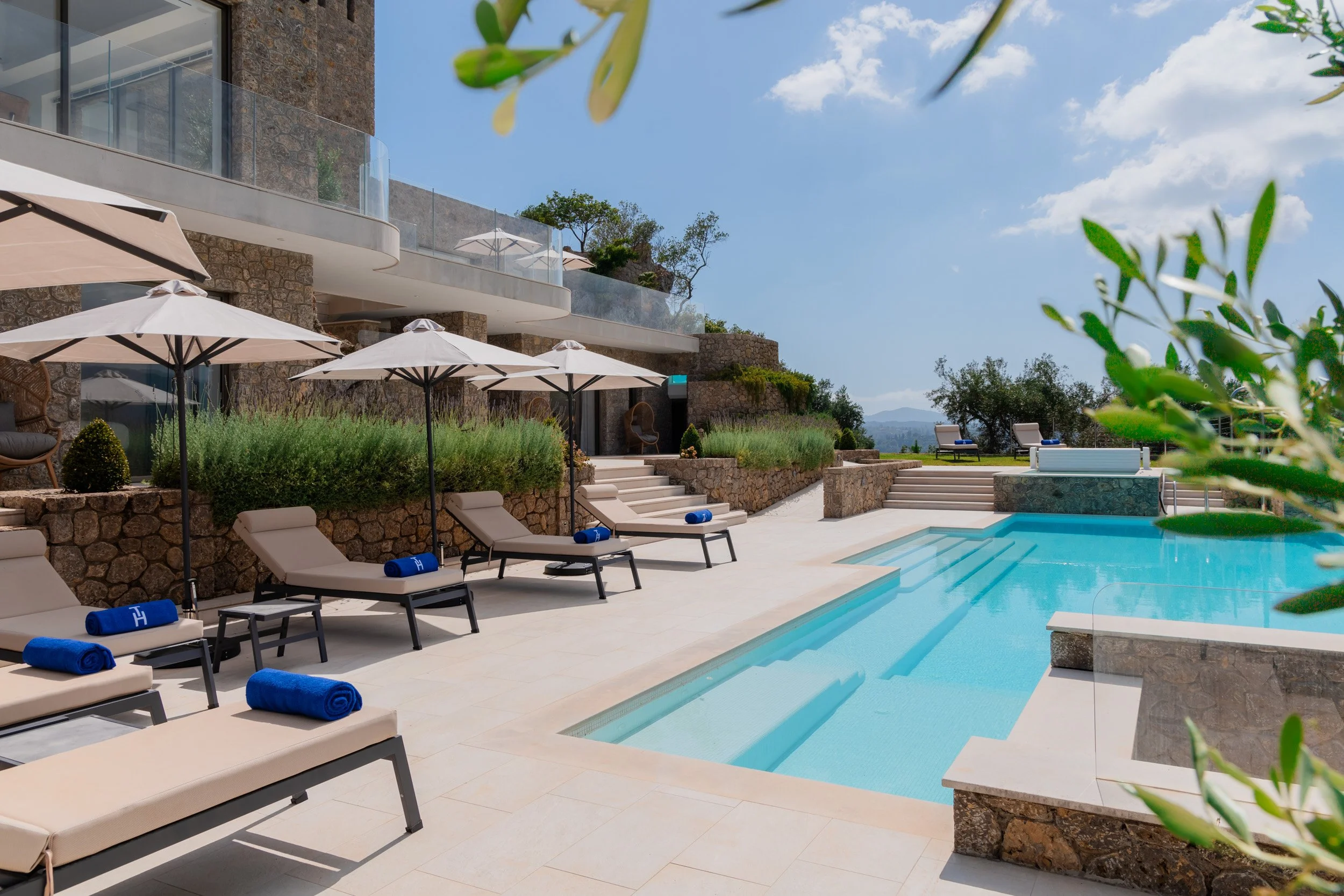 Luxury outdoor swimming pool area with beige lounge chairs, blue rolled towels, white umbrellas, stone building, and hillside landscape under a blue sky with clouds.