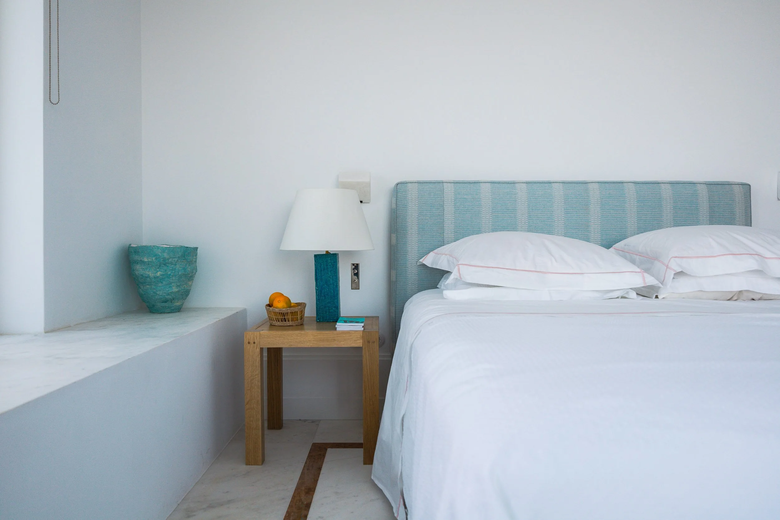 A neatly made white bed with pillows, a wooden nightstand with a lamp, a basket of oranges, and a small stack of books, in a minimalist room with white walls and a marble ledge.