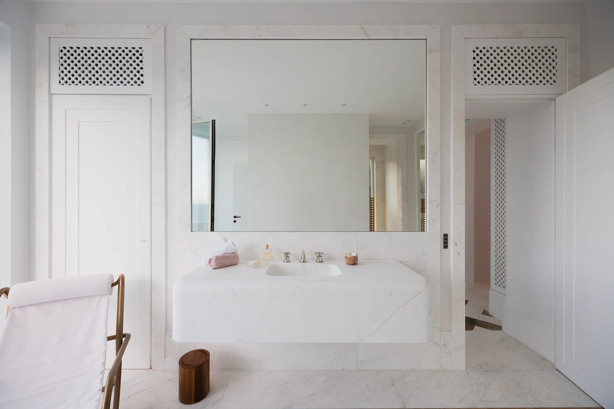 A white bathroom with a large mirror above a marble sink, towels, soap, and a small container on the countertop, with white walls and minimal decor.