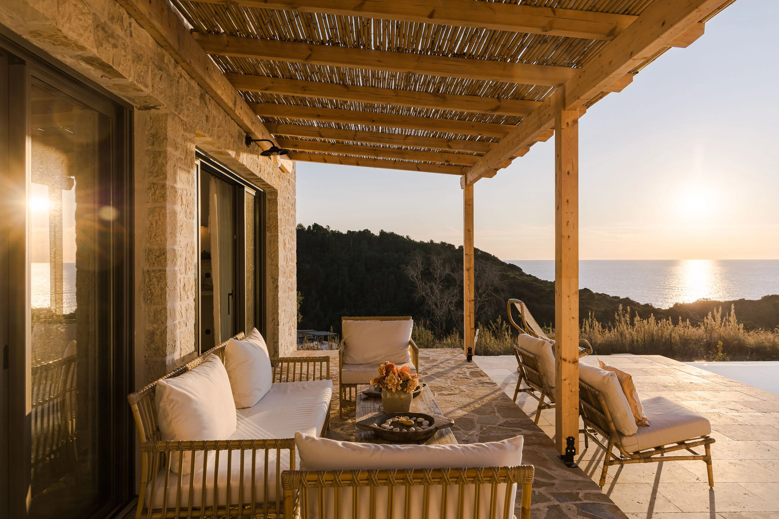 Outdoor patio with wicker furniture, cushions, and a flower arrangement, overlooking a sunset over hillside and ocean.