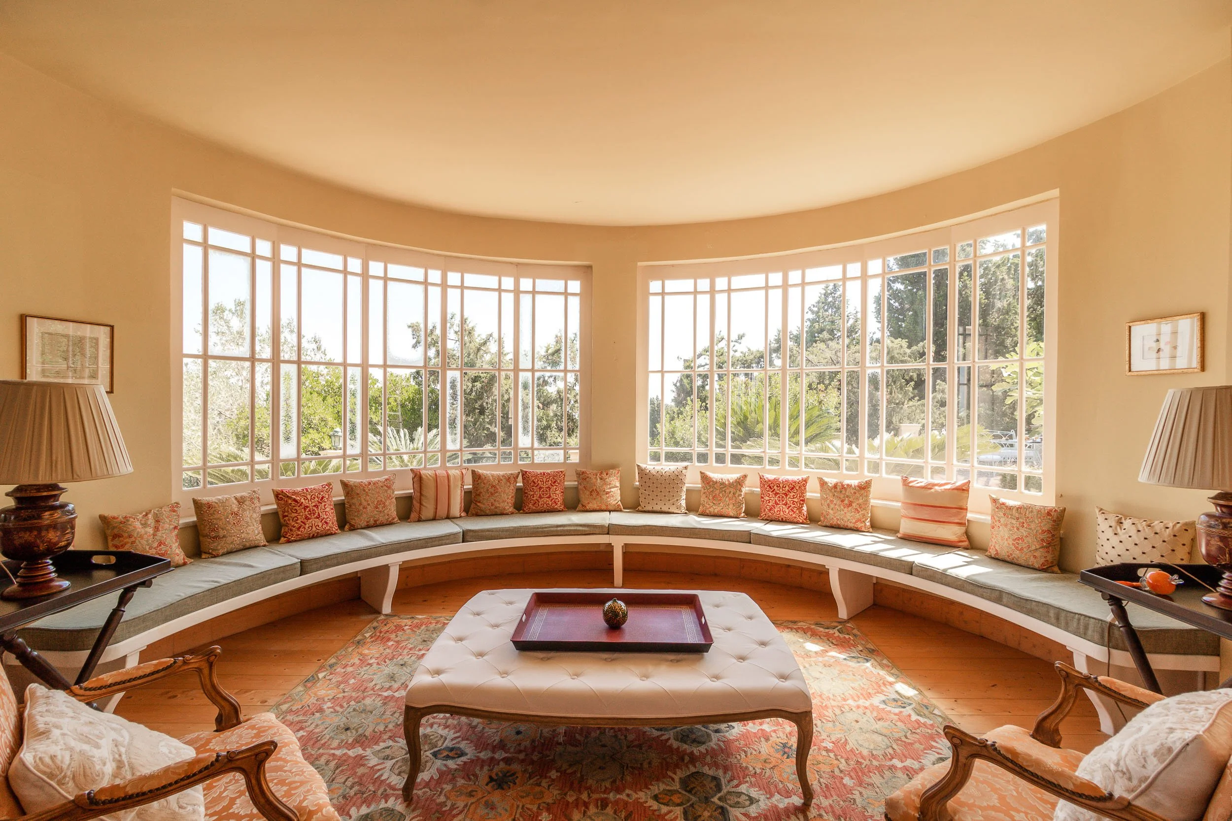 Bright living room with a large curved window, beige walls, and a patterned area rug. Contains a tufted ottoman with a tray and a decorative ball, cushioned built-in window seat with multiple pillows, armchairs, and side tables with lamps.