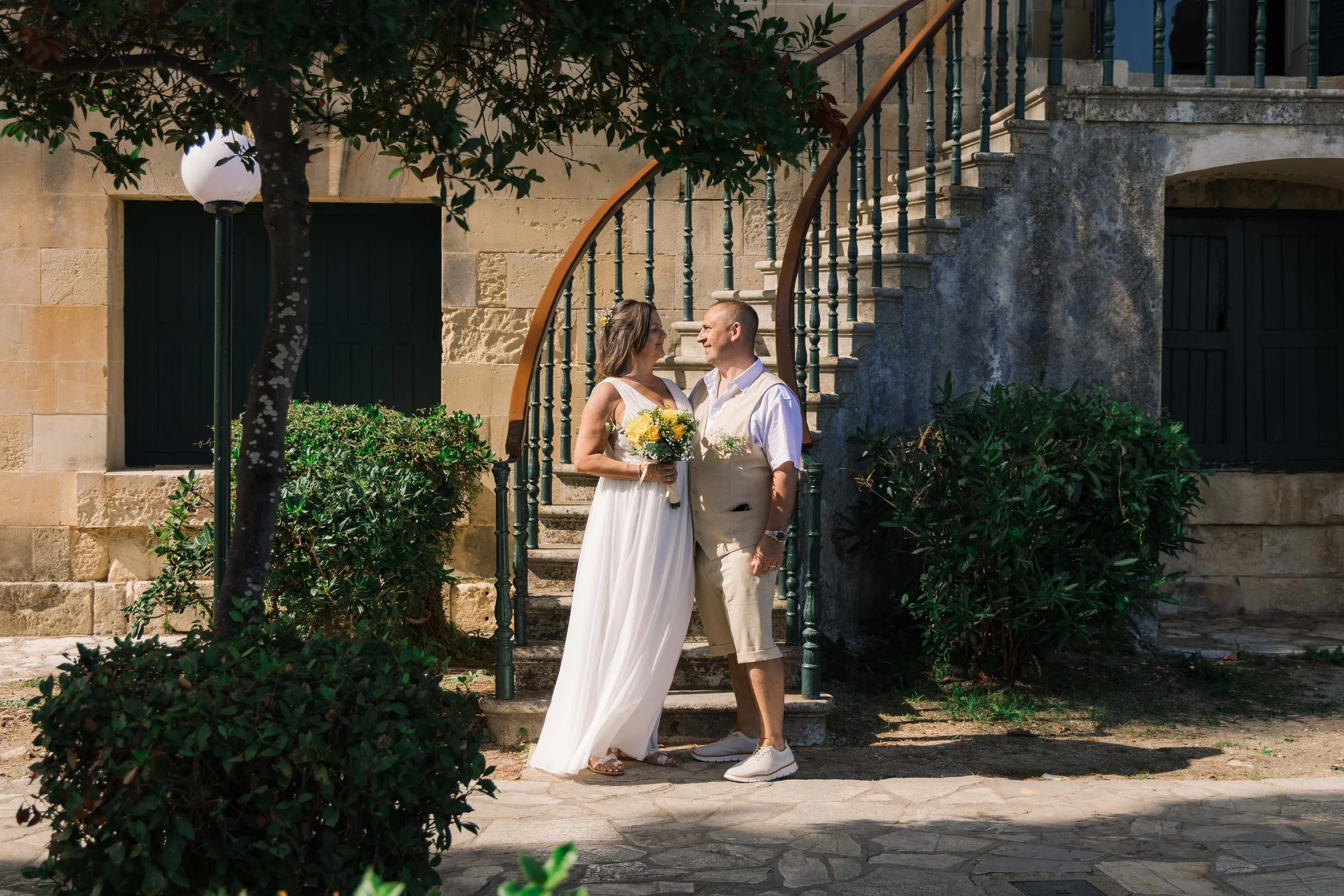 A couple dressed in wedding attire standing close together outdoors near stone stairs, surrounded by green bushes and trees, with a stone building and a lamp post in the background.