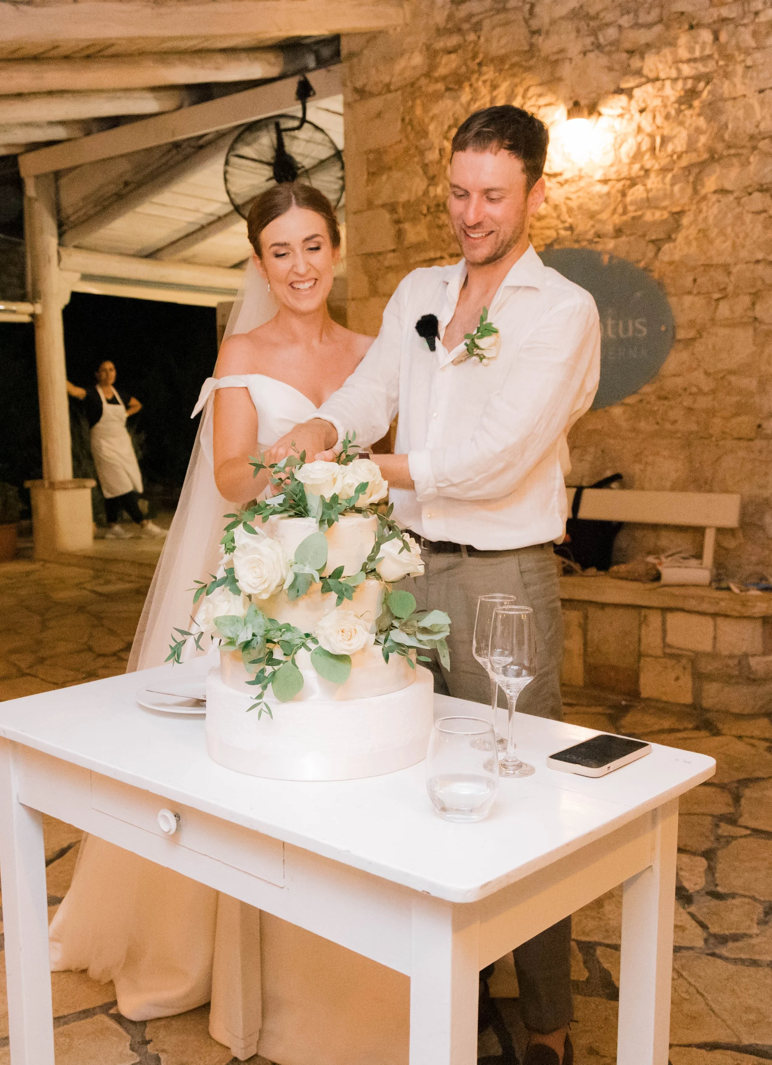 A bride and groom cutting a wedding cake together at their reception. The three-tiered cake is decorated with white flowers and greenery. The couple is smiling and dressed in wedding attire. The setting is a rustic venue with exposed brick walls.